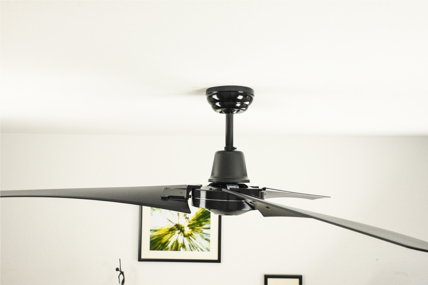 Schwarzer Deckenventilator mit vier Flügeln, an einer weißen Decke montiert. Ein gerahmtes Bild ist sichtbar.