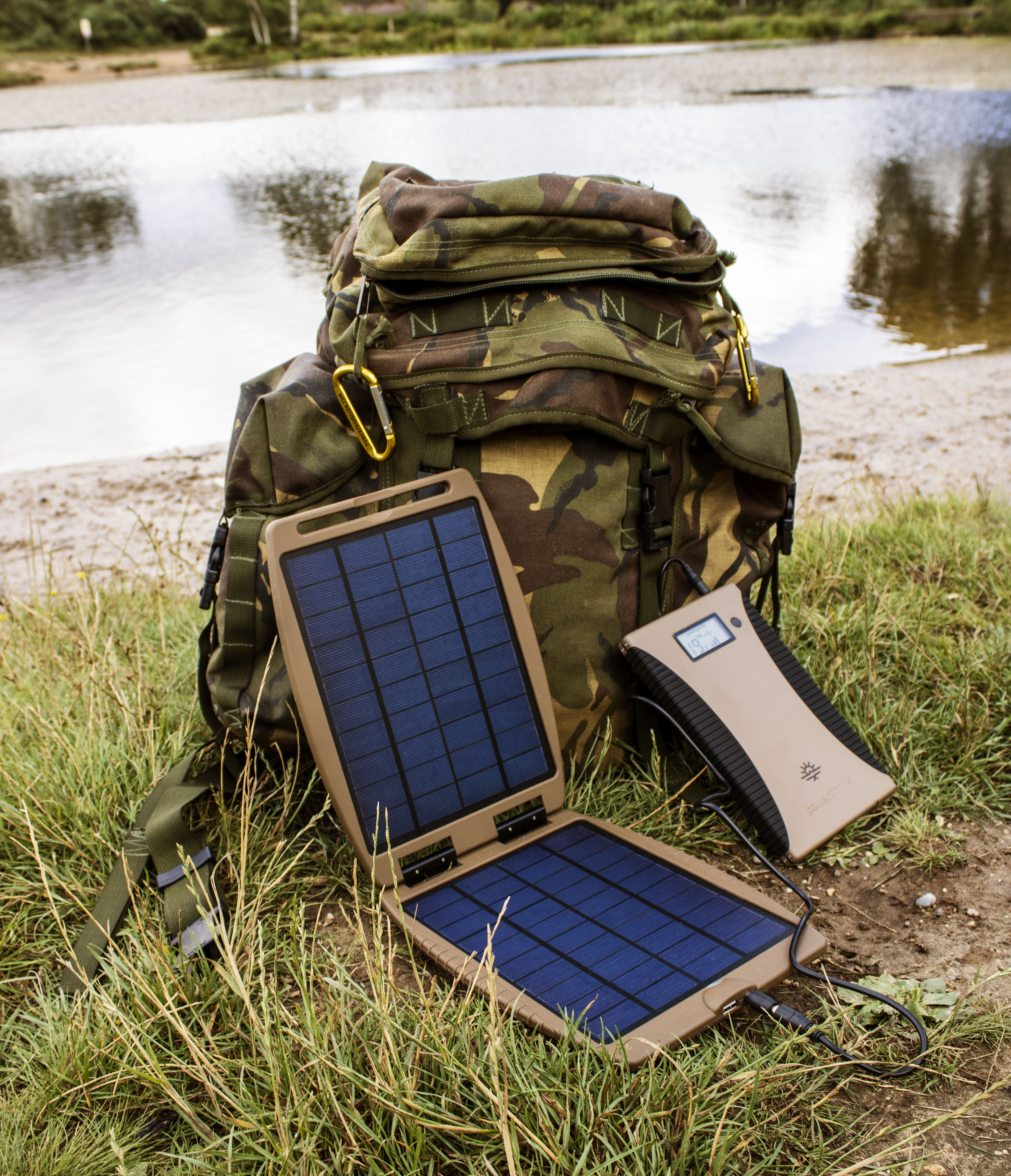 Ein Camouflage-Rucksack neben einem Solarladegerät und einer Powerbank auf Gras am Wasser.