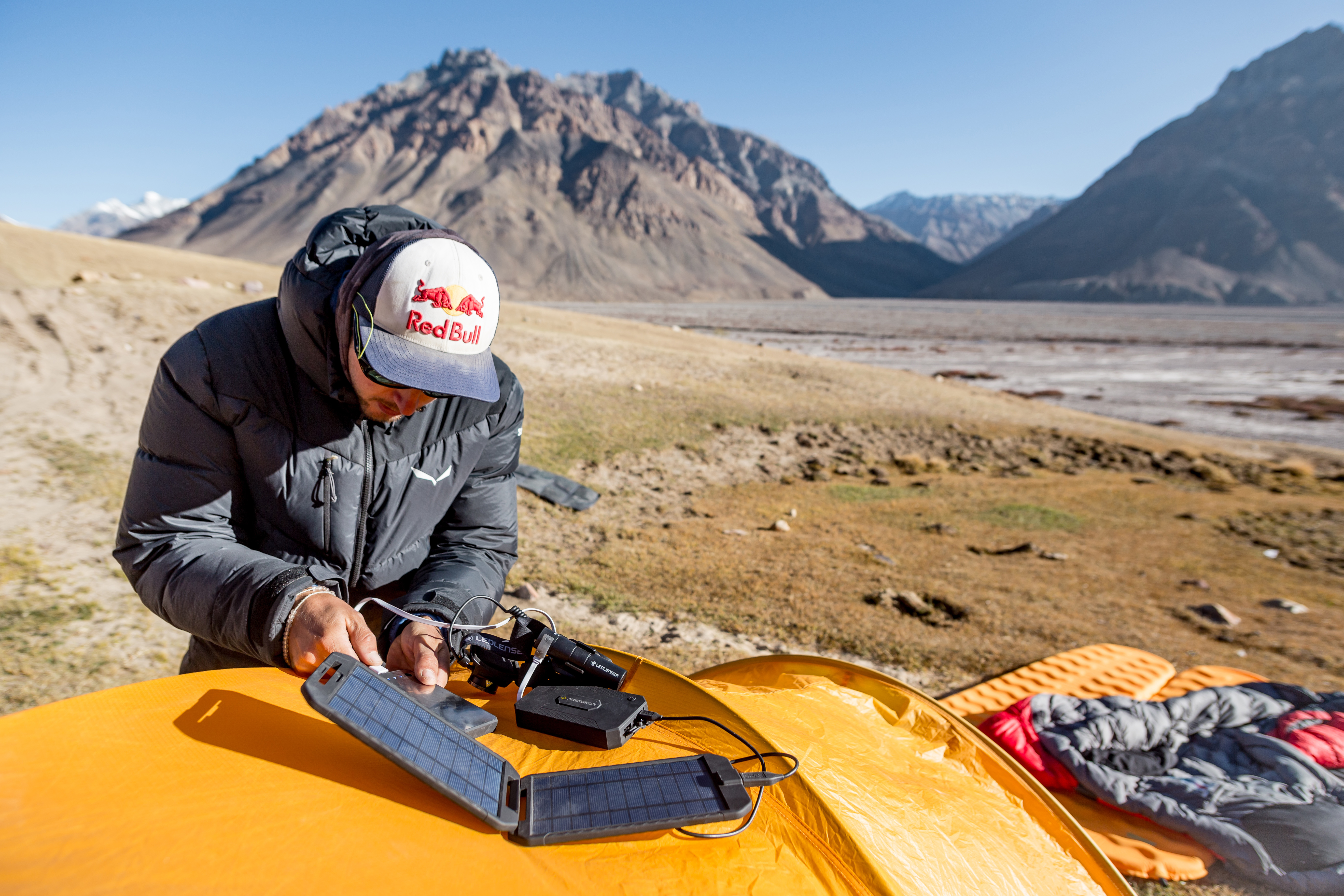 Mann mit Hut arbeitet mit Solarpanel, in der Nähe eines Zeltes, in Berglandschaft.