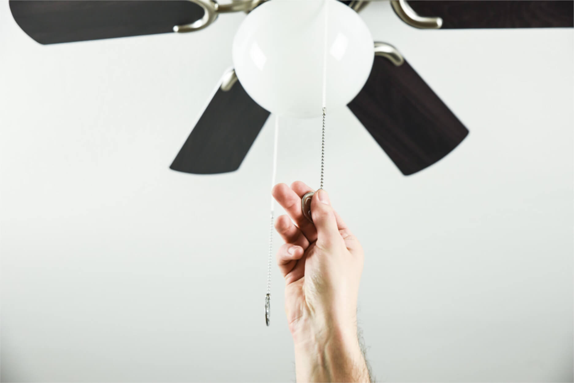 Hand zieht Kette an Deckenventilator mit Leuchte vor weißer Wand.
