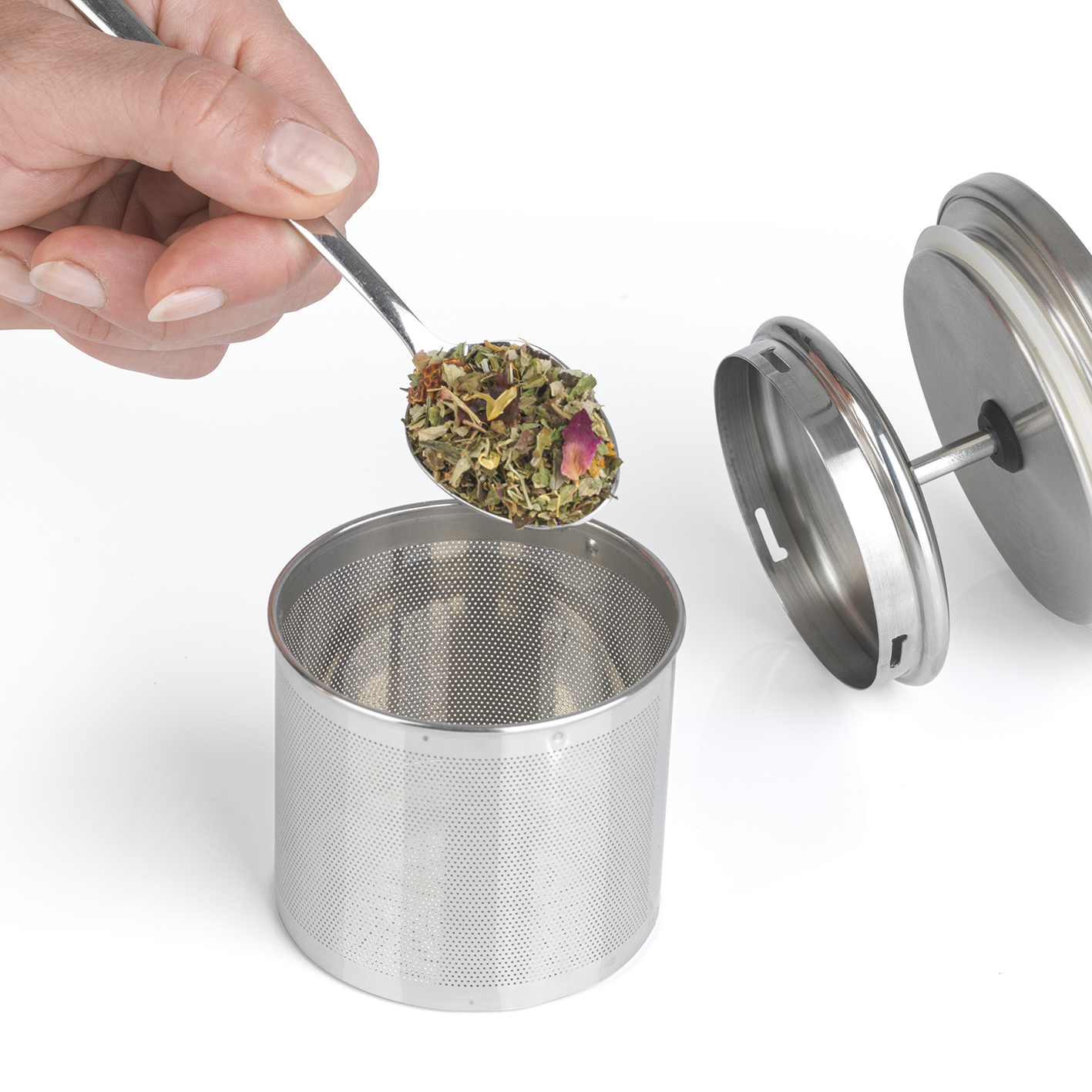 Eine Hand gießt mit einem Löffel Teeblätter in einen Edelstahl-Teezubereiter, mit offenem Deckel.