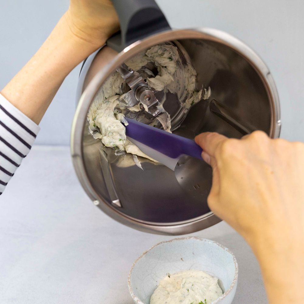 Mani che usano una spatola viola per rimuovere il contenuto da una ciotola d'argento di un robot da cucina in una ciotola bianca.
