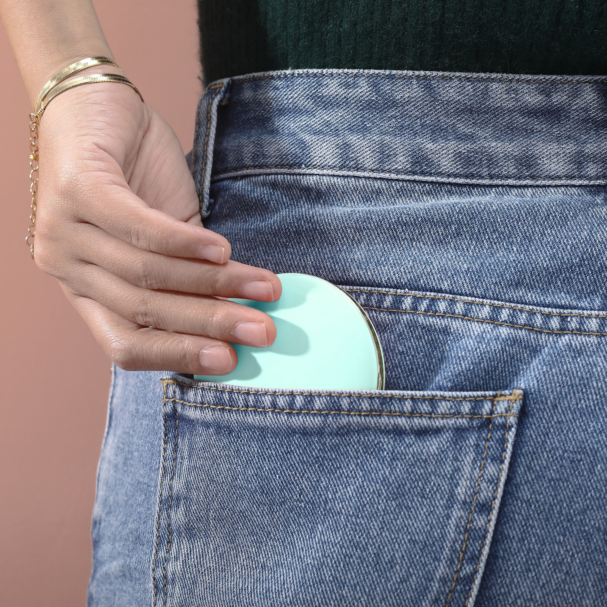 Eine Hand steckt einen kleinen, runden, mintgrünen Kompaktspiegel in die Gesäßtasche einer Jeans.