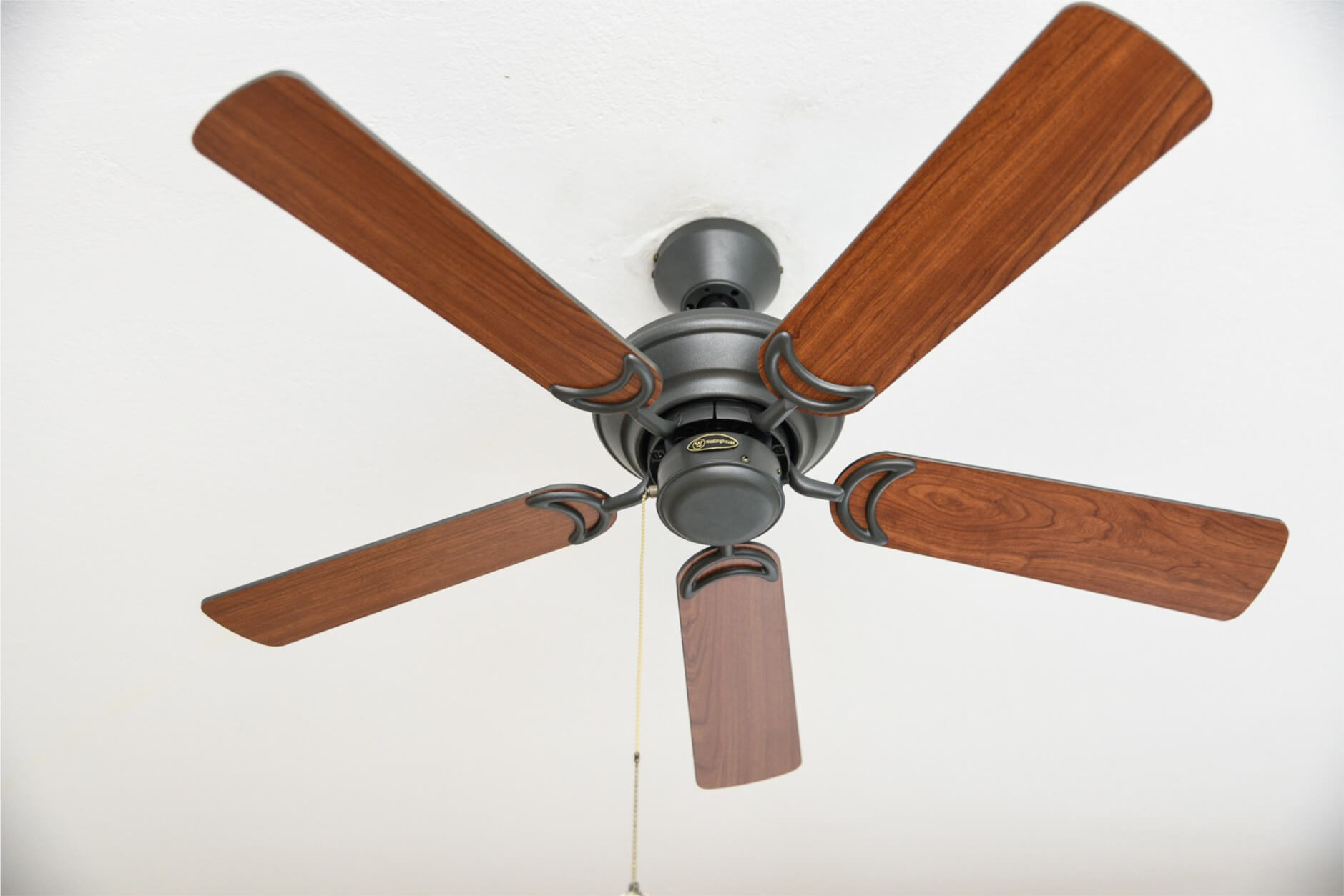 Ein Deckenventilator mit fünf Holzflügeln und einem dunkelgrauen Motor vor weißer Decke.