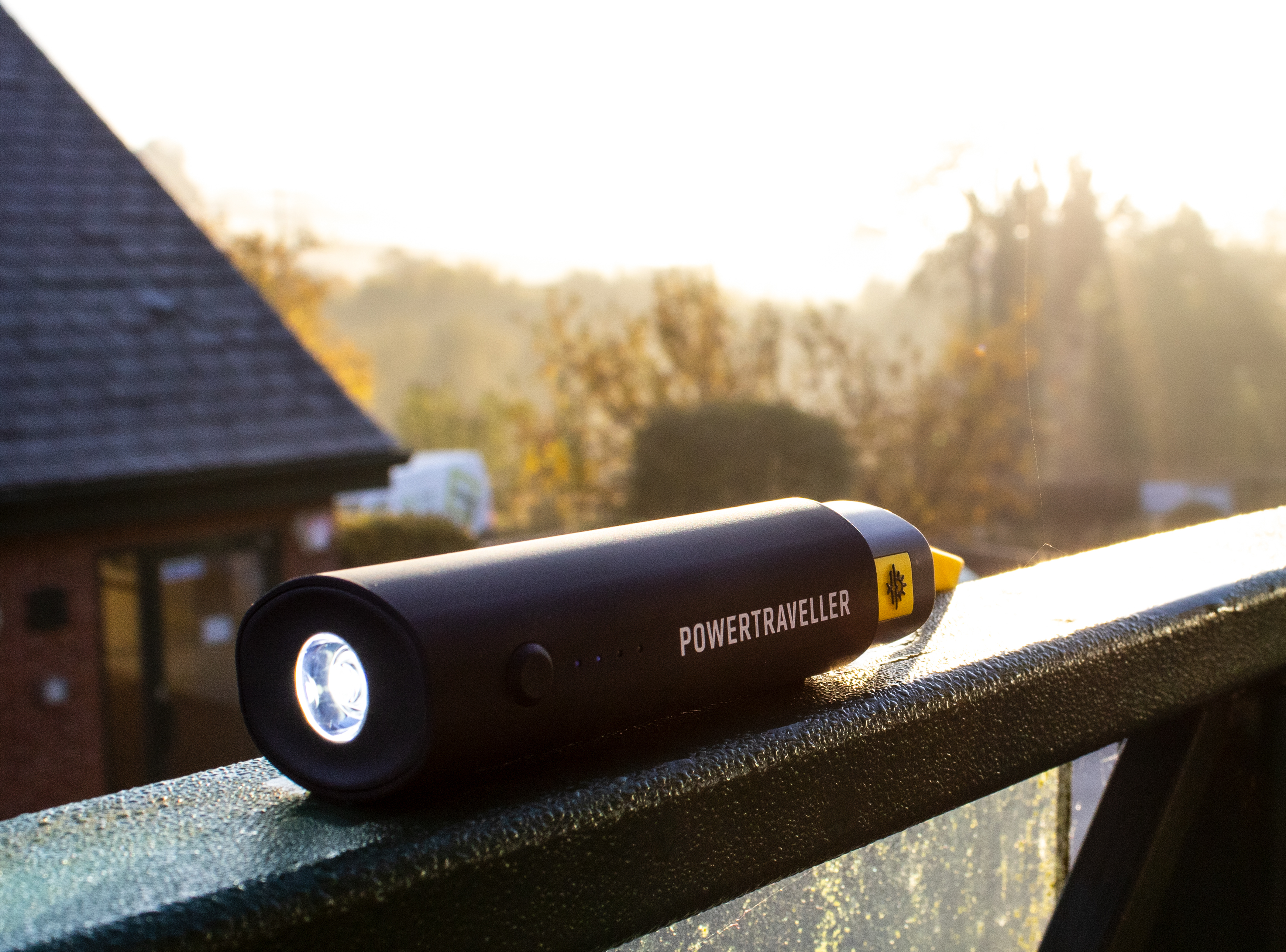 Schwarze, zylindrische Taschenlampe mit Licht. Draußen auf einem Geländer. Bewölkter Himmel und Bäume im Hintergrund.