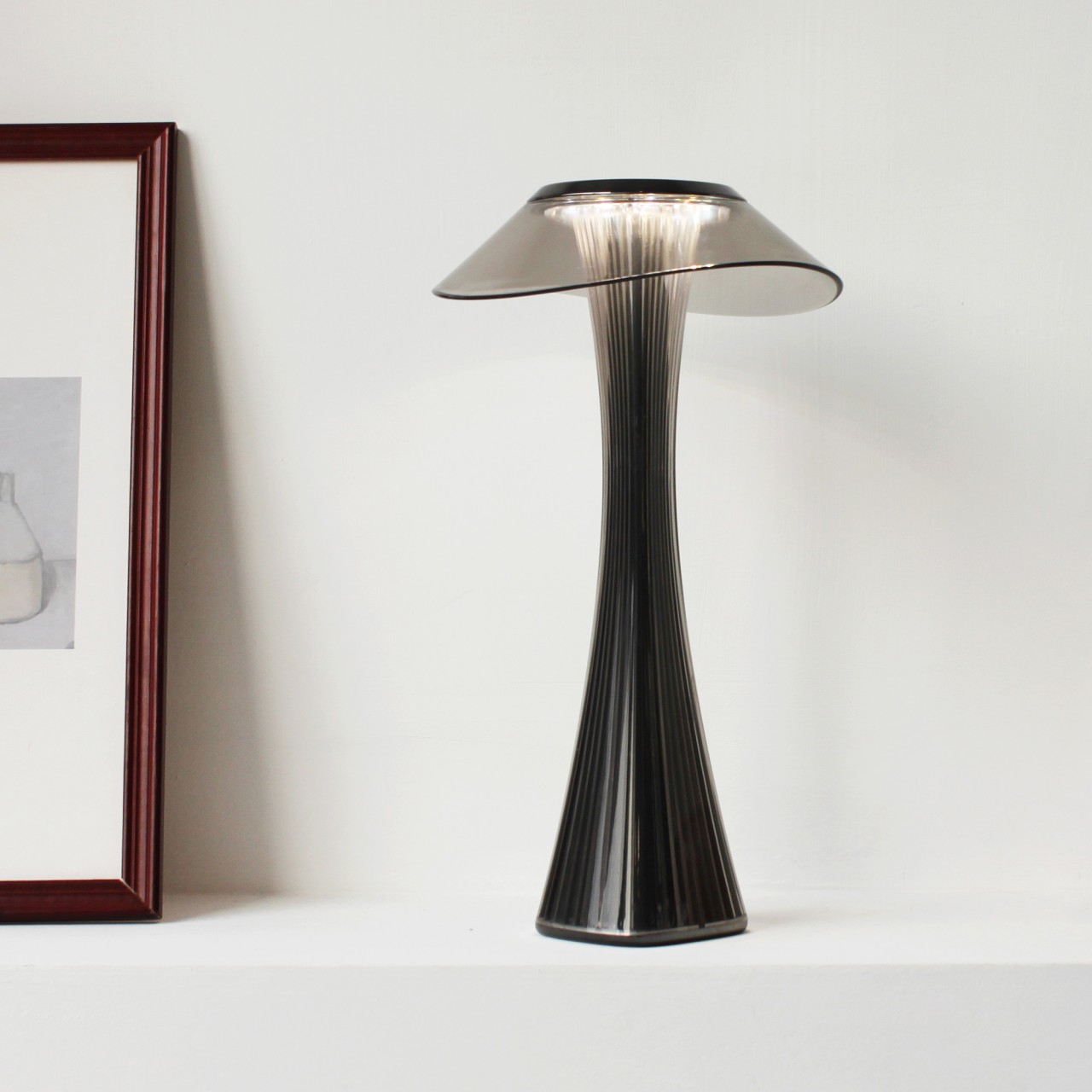Eine schwarz-silberne Lampe auf einem weißen Regal, mit einem gerahmten Bild im Hintergrund.