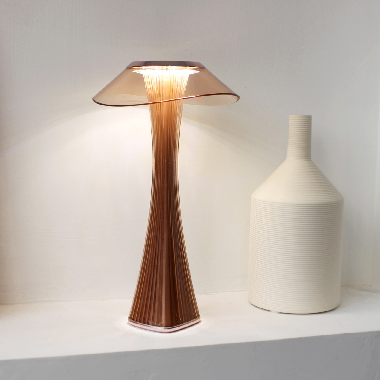 Moderne braune Lampe und eine weiße Vase auf einem weißen Regal. Die Lampe ist beleuchtet.