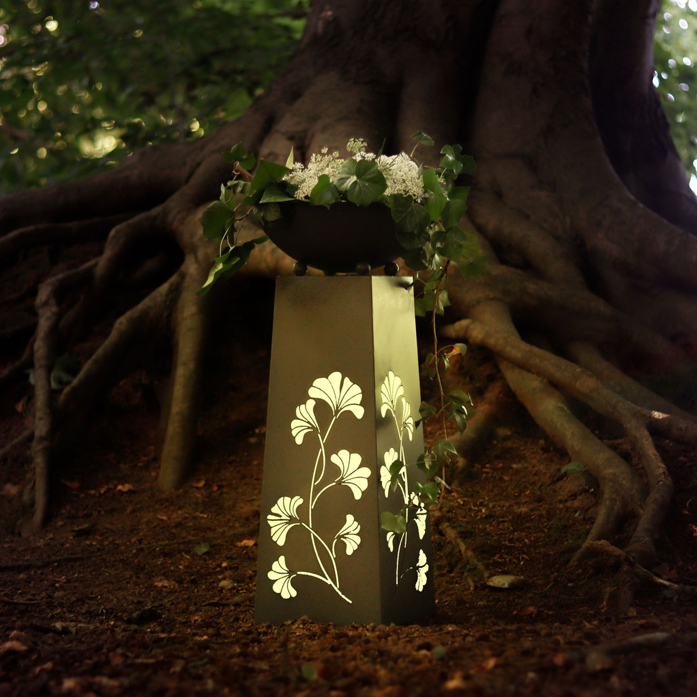 Ein schwarzer, blumengeschmückter Sockel mit einer Schale mit Pflanzen im Wald.