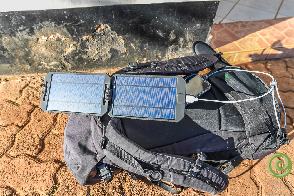 Schwarzer Rucksack auf Ziegeloberfläche, mit offenem Solarpanel, lädt Telefon per USB-Kabel.
