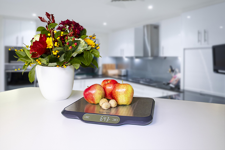 Waga kuchenna z jabłkami, orzechami włoskimi i kwiatami. Waga pokazuje 643 gramy. Biała kuchnia w tle.