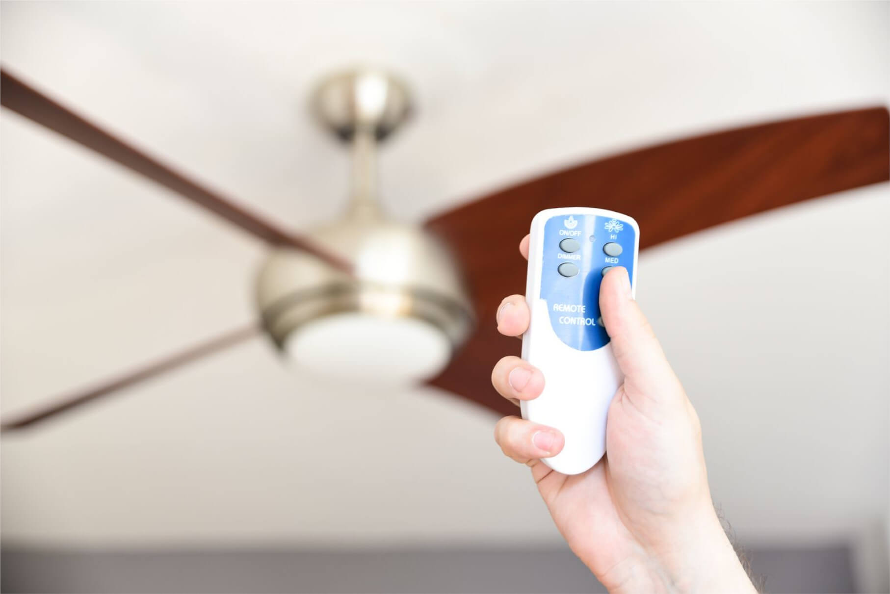 Hand hält Fernbedienung, Deckenventilator im Hintergrund. Lüfterflügel sind dunkelbraun.