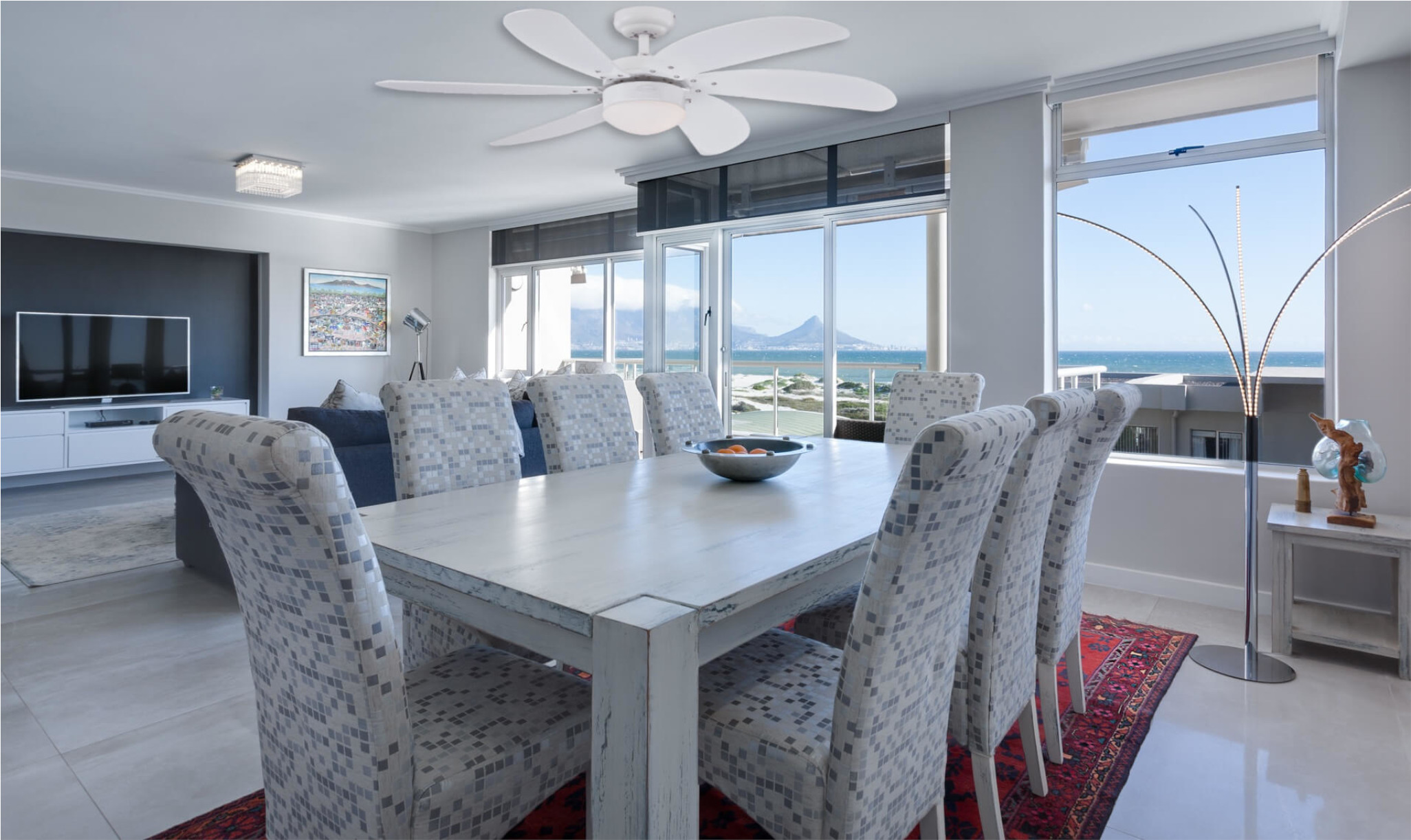 Esszimmer mit Tisch, Stühlen, Ventilator und Blick auf Meer und Berge durch Fenster. Eine Schüssel mit Orangen steht auf dem Tisch.