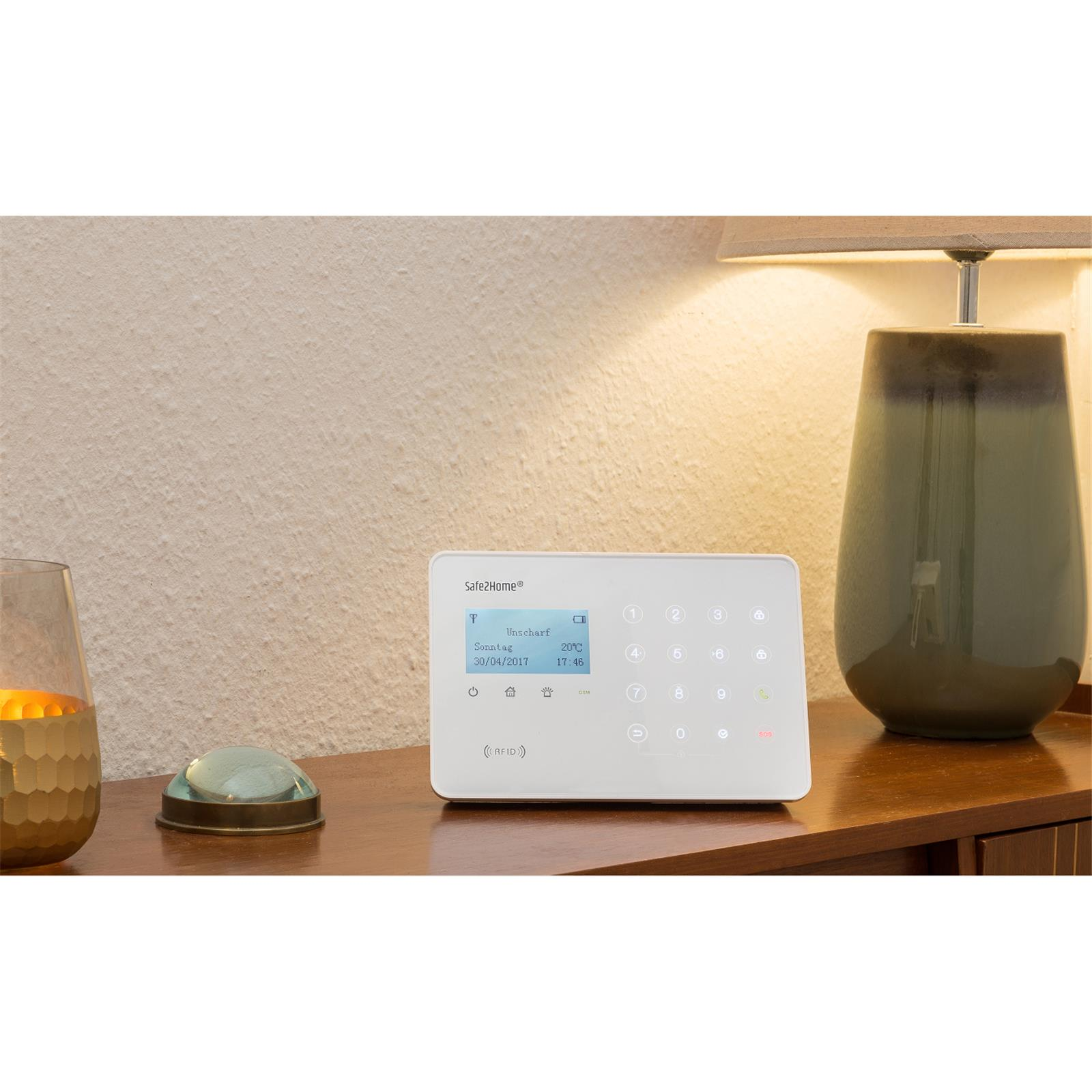 Ein weißes Sicherheitssystem-Panel steht auf einer Holzoberfläche mit einer Lampe und einem dekorativen Gegenstand.