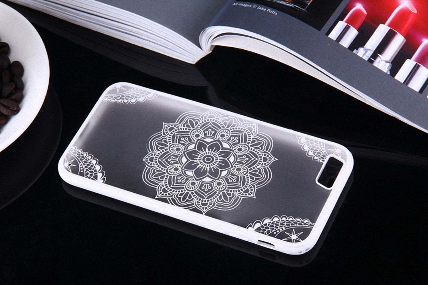 Transparente Handyhülle mit weißem Mandala-Design auf schwarzem Untergrund mit offenem Buch und Kaffee.