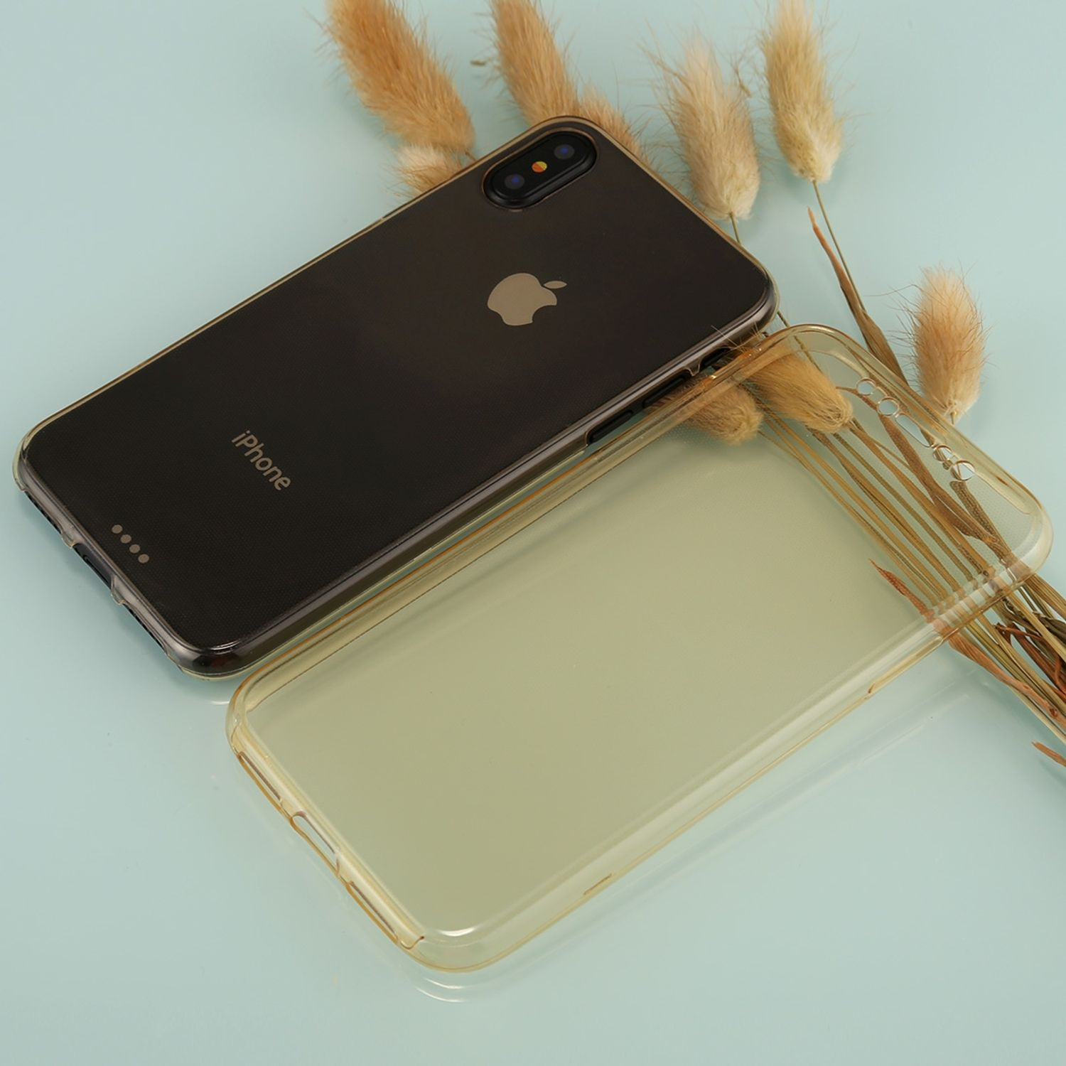 Schwarzes iPhone mit klarer Hülle, plus eine klare Hülle an der Seite, mit einigen trockenen Pflanzen im Hintergrund.