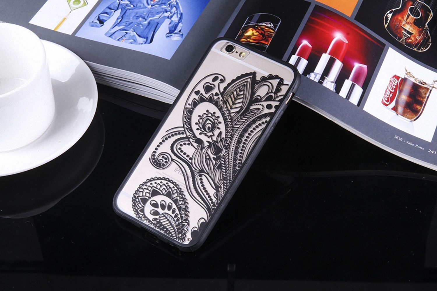 Ein Smartphone mit einer schwarzen gemusterten Hülle liegt auf einer reflektierenden schwarzen Oberfläche neben einer Tasse und einer Zeitschrift.