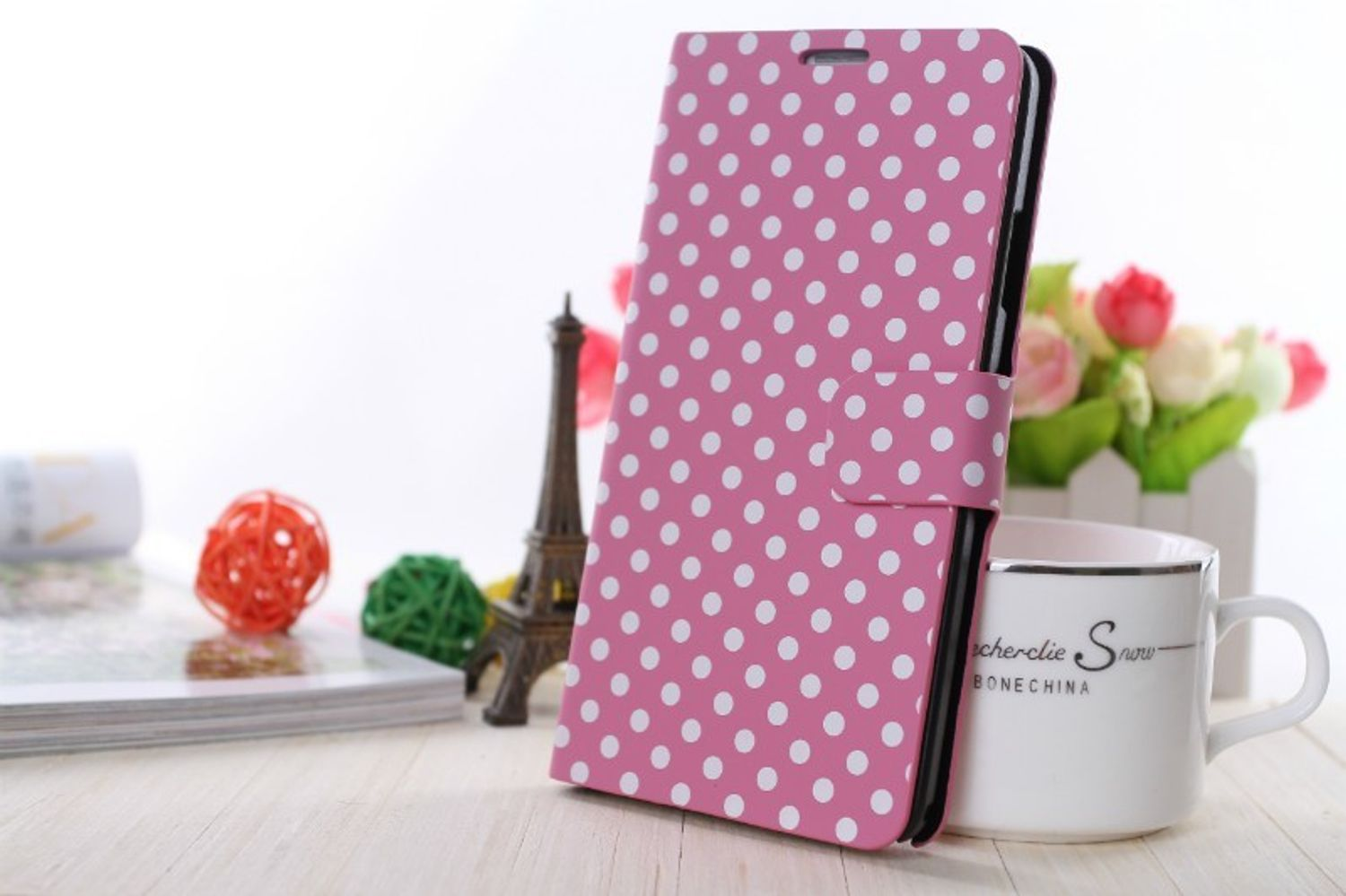 Rosa Polka Dot Handyhülle mit weißem Hintergrund. Eine Tasse, Blumen und andere Dekorationen sind im Hintergrund.