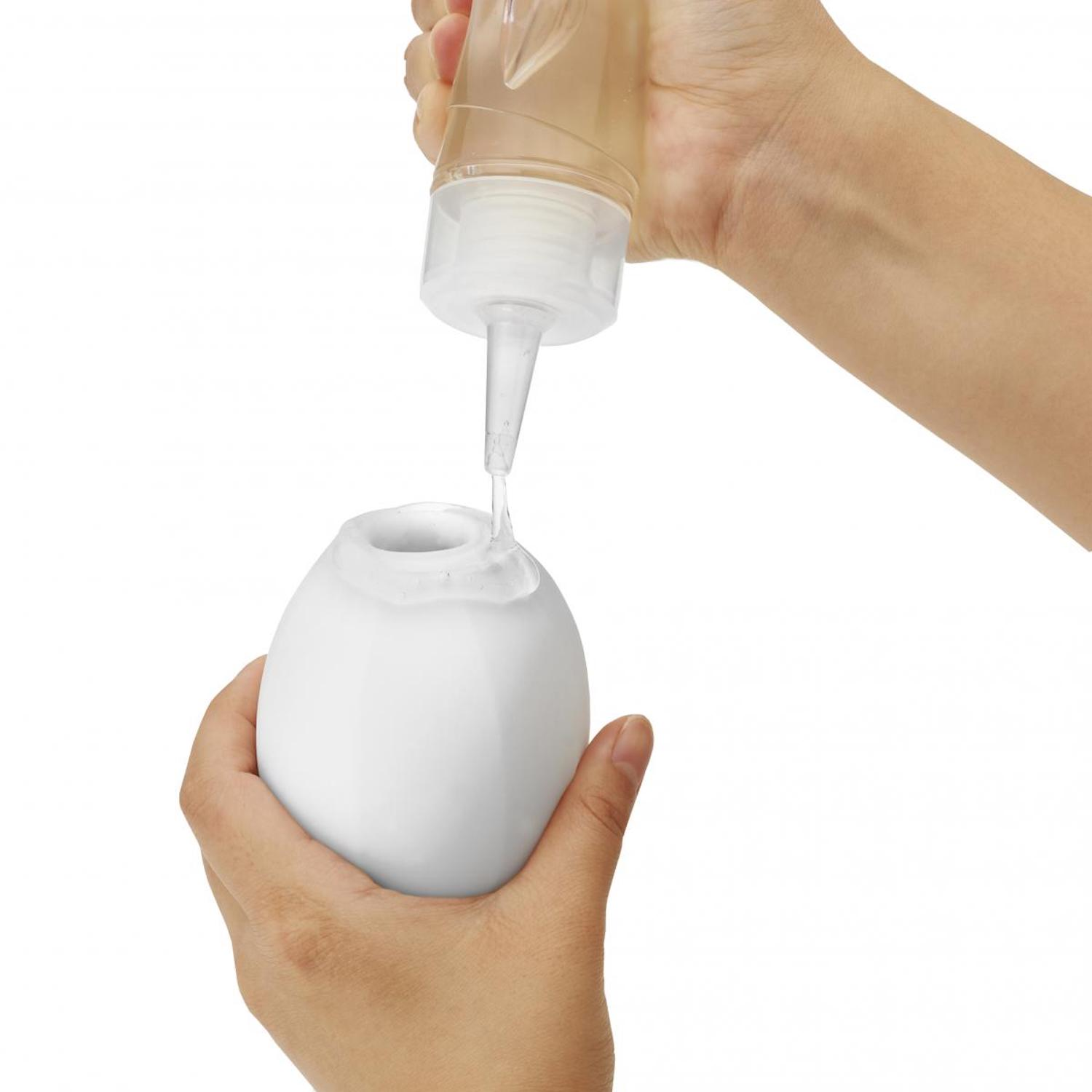 Eine Person gießt Flüssigkeit aus einer Flasche in einen weißen, eiförmigen Behälter. Weißer Hintergrund.