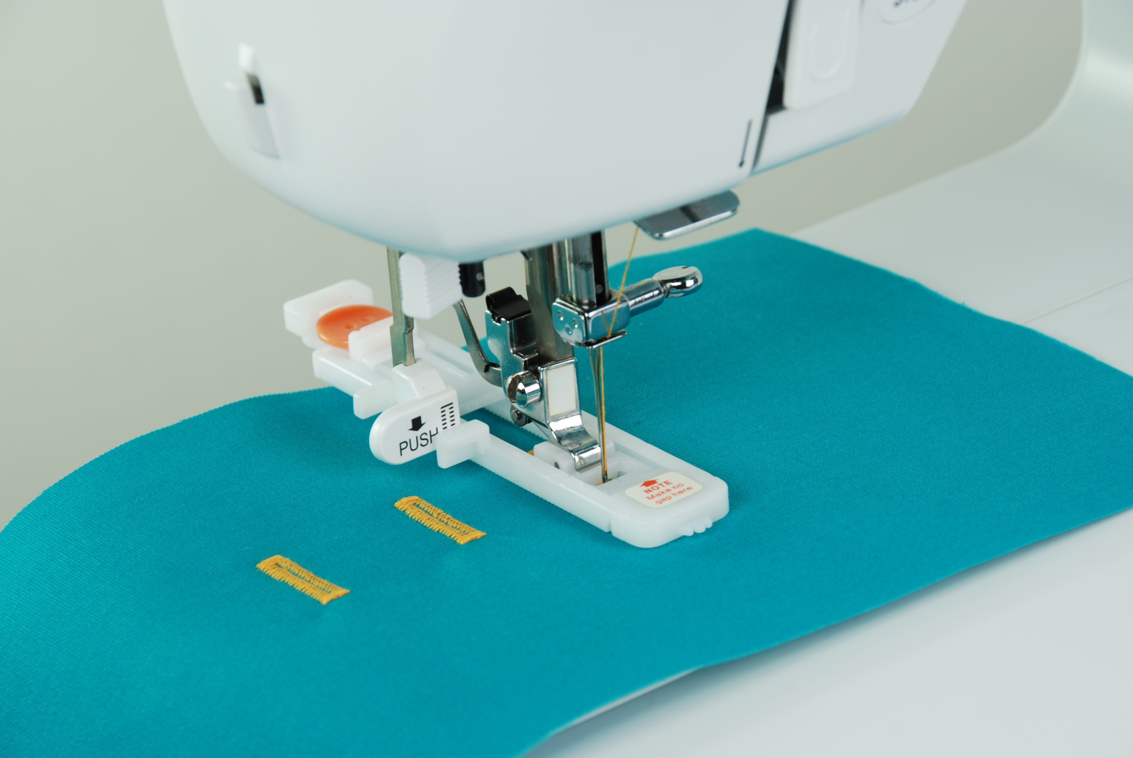 Máquina de coser cosiendo sobre tela azul. La máquina es blanca y tiene guías para ojales amarillas.