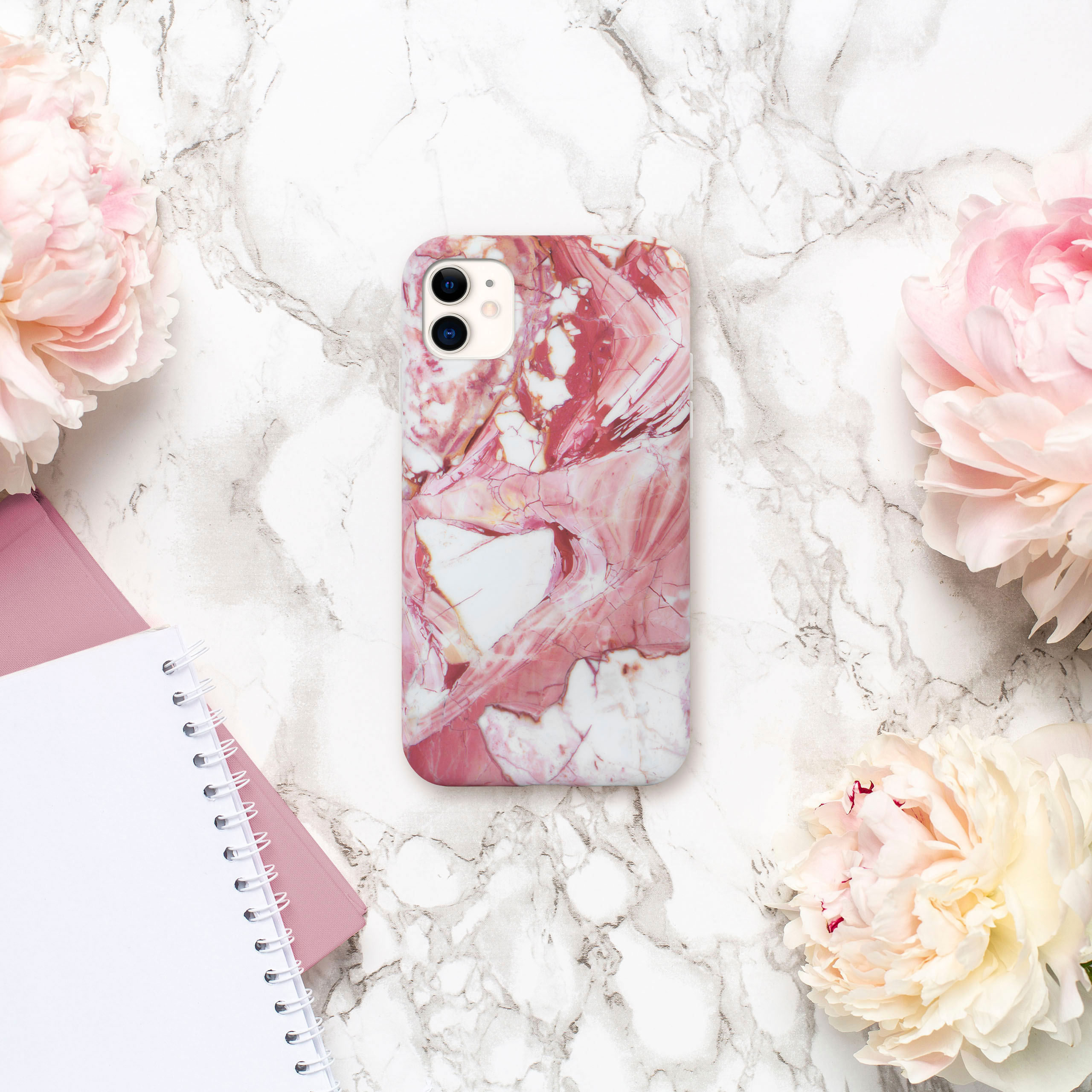 Handyhülle mit Marmordesign, umgeben von rosa Blumen und einem Notizblock. Weißer Hintergrund.
