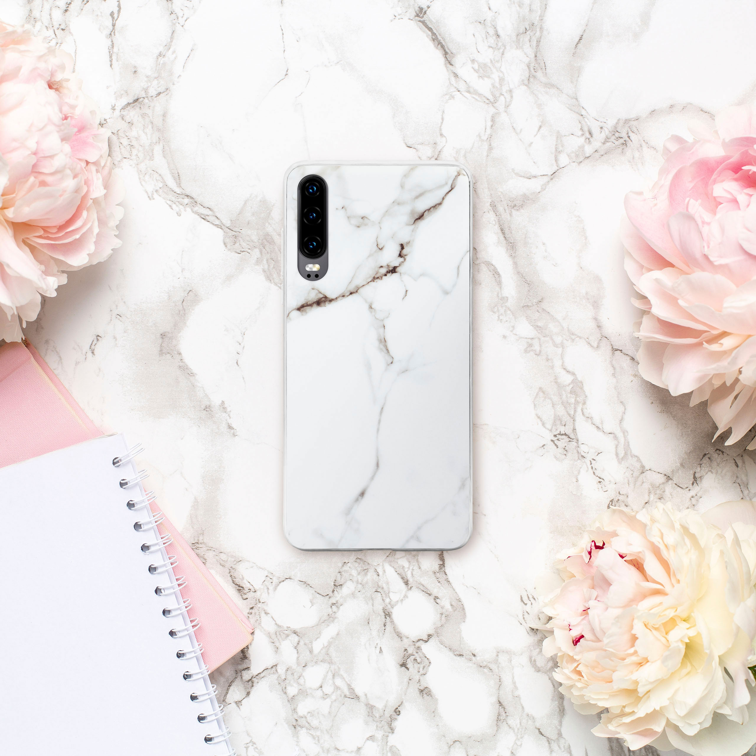 Handyhülle mit weißem Marmordesign, rosa Notizbuch und Blumen auf Marmoroberfläche.