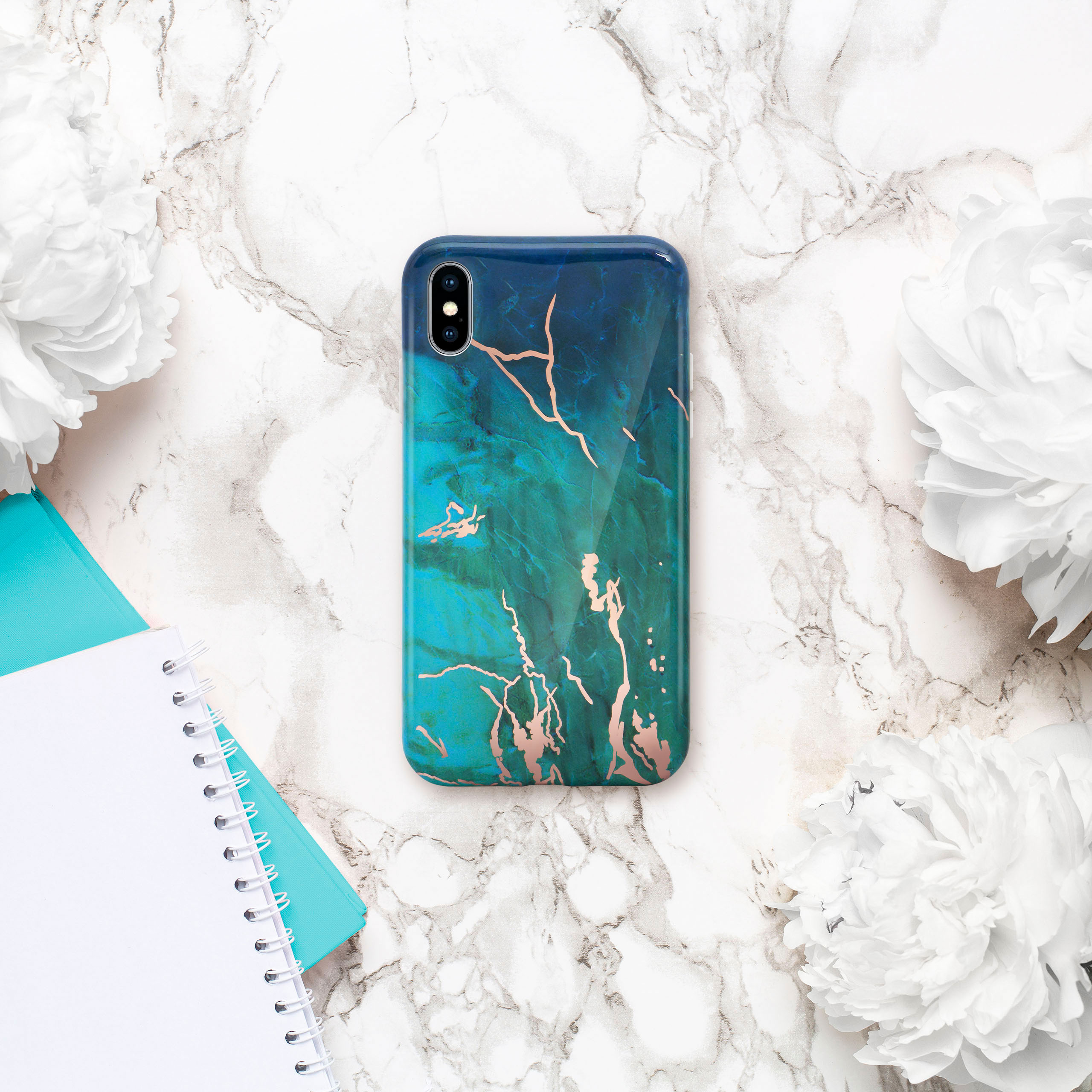 Handyhülle mit blauem und goldenem Marmormuster. Daneben Blumen und ein Notizbuch auf Marmor.