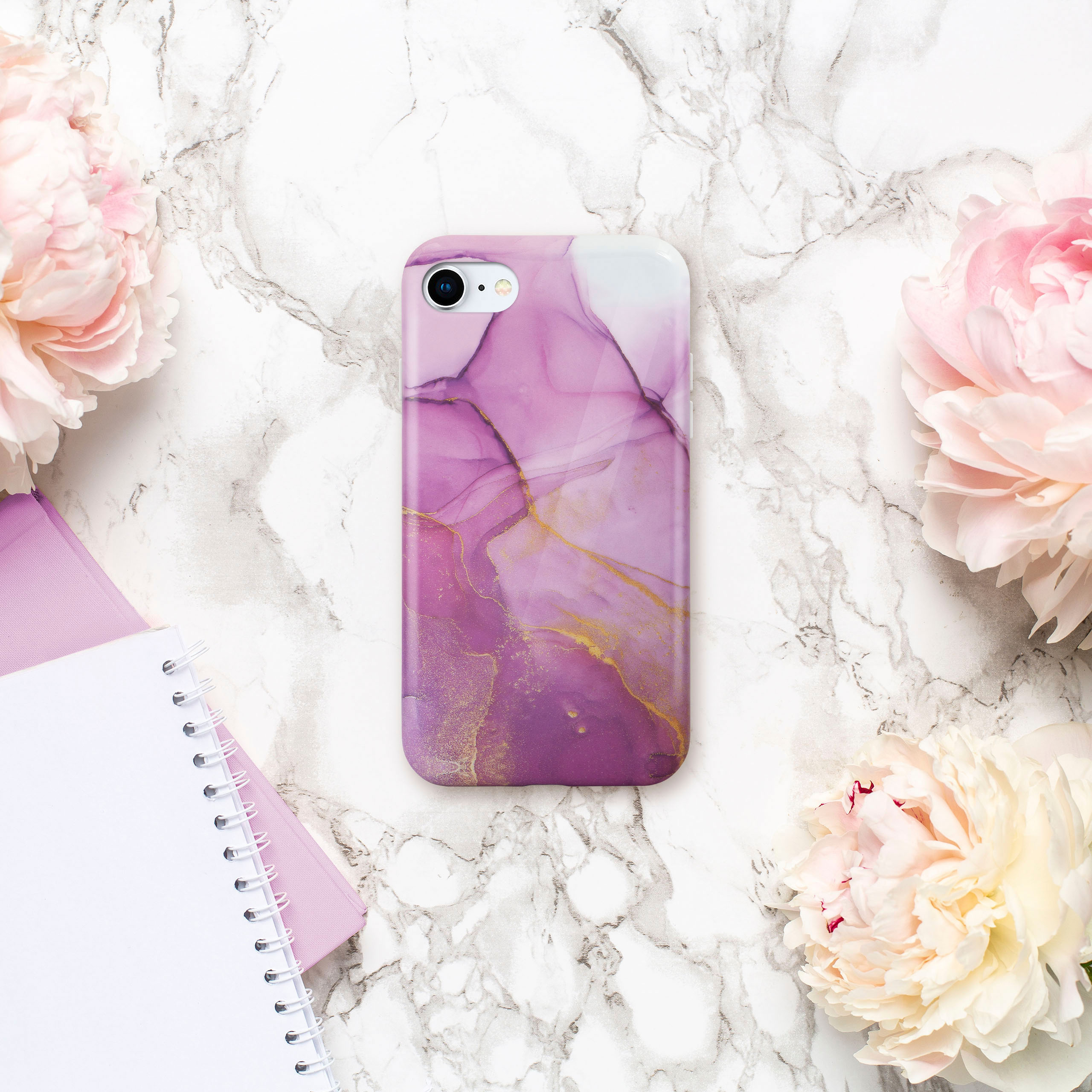 Handyhülle mit lila-goldenem, abstraktem Design liegt zwischen Blumen und Notizbüchern.