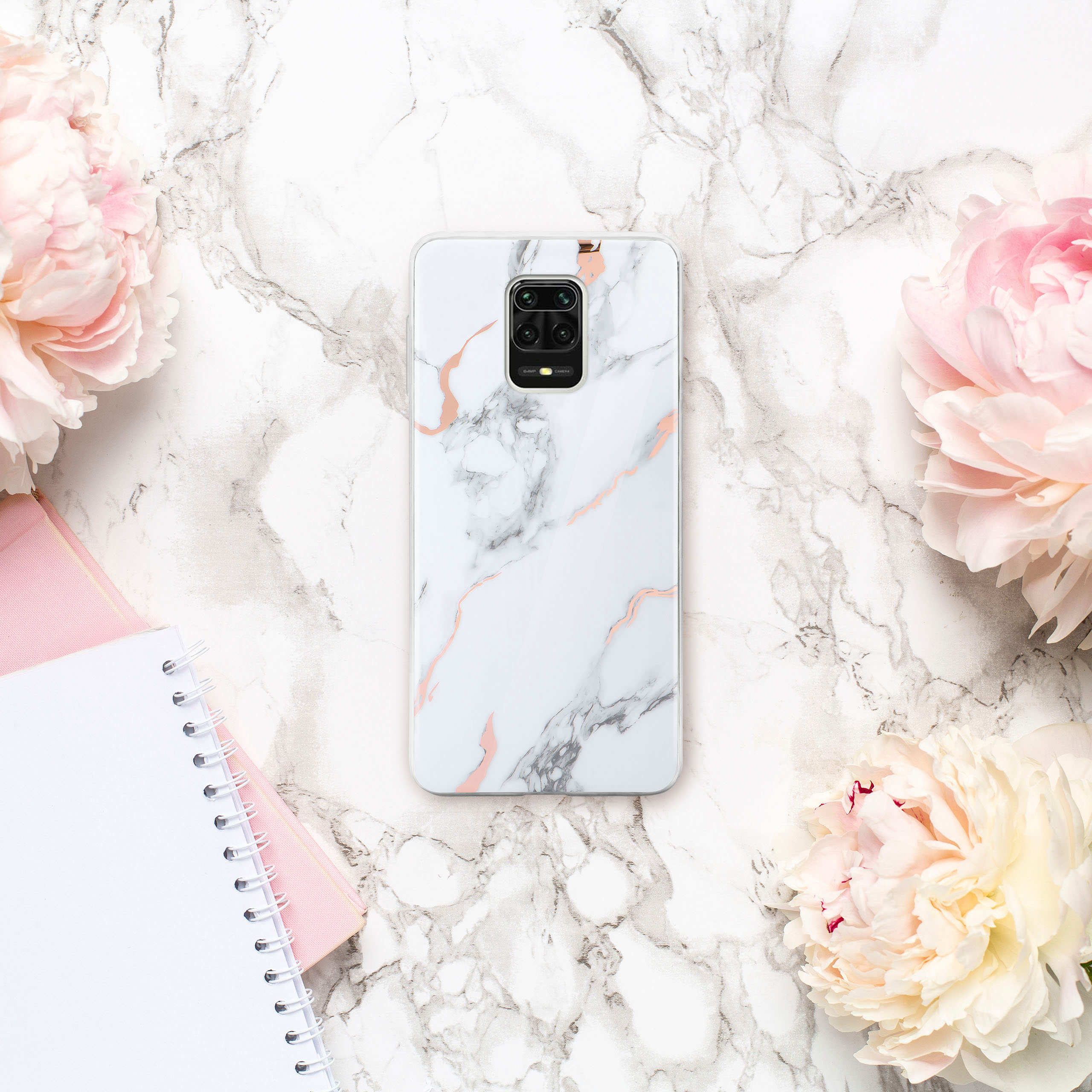 Handyhülle mit Marmordesign, umgeben von Blumen und Notizbüchern auf einer Marmoroberfläche.