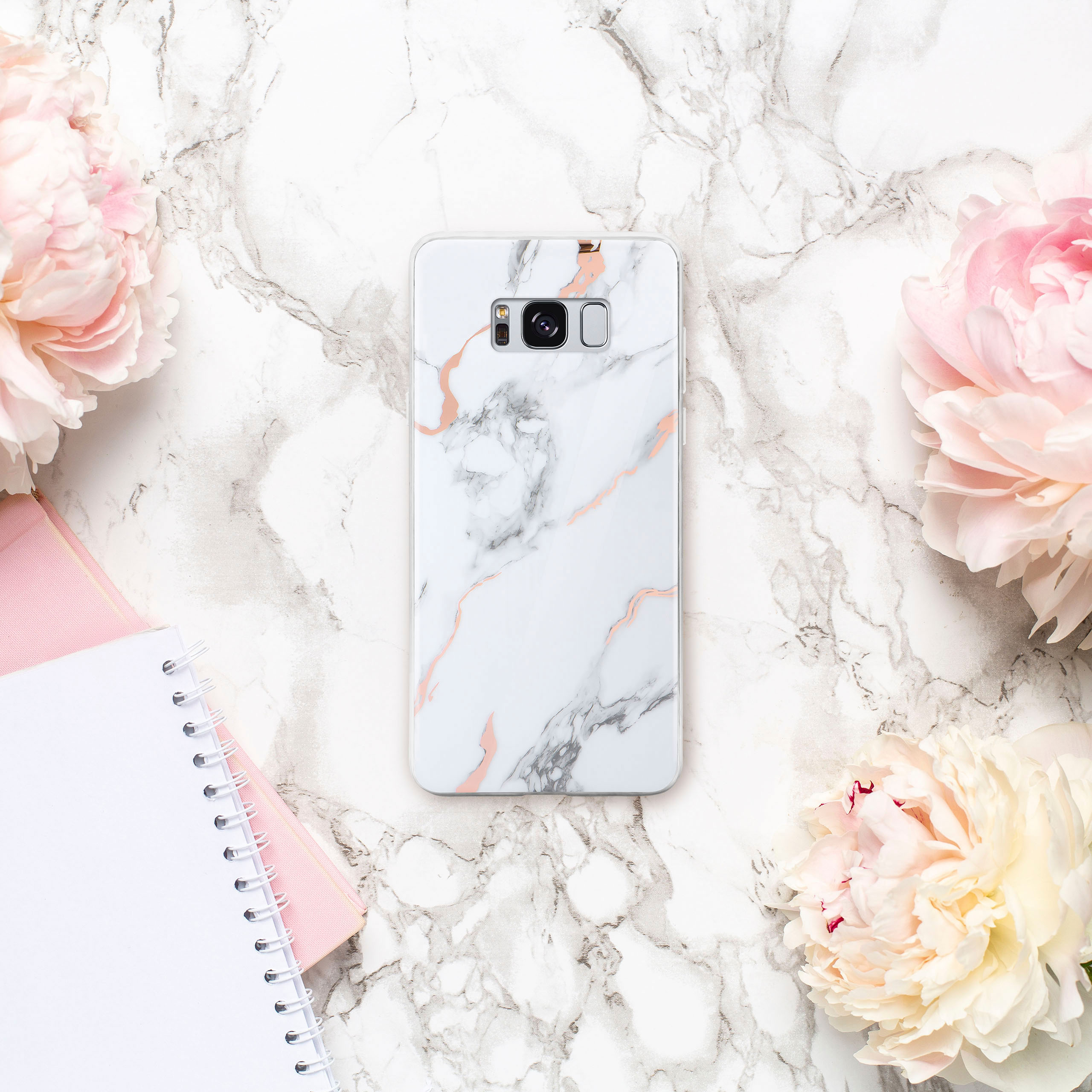 Handyhülle mit Marmor-Design, rosa Blumen und Notizbuch auf Marmorhintergrund.