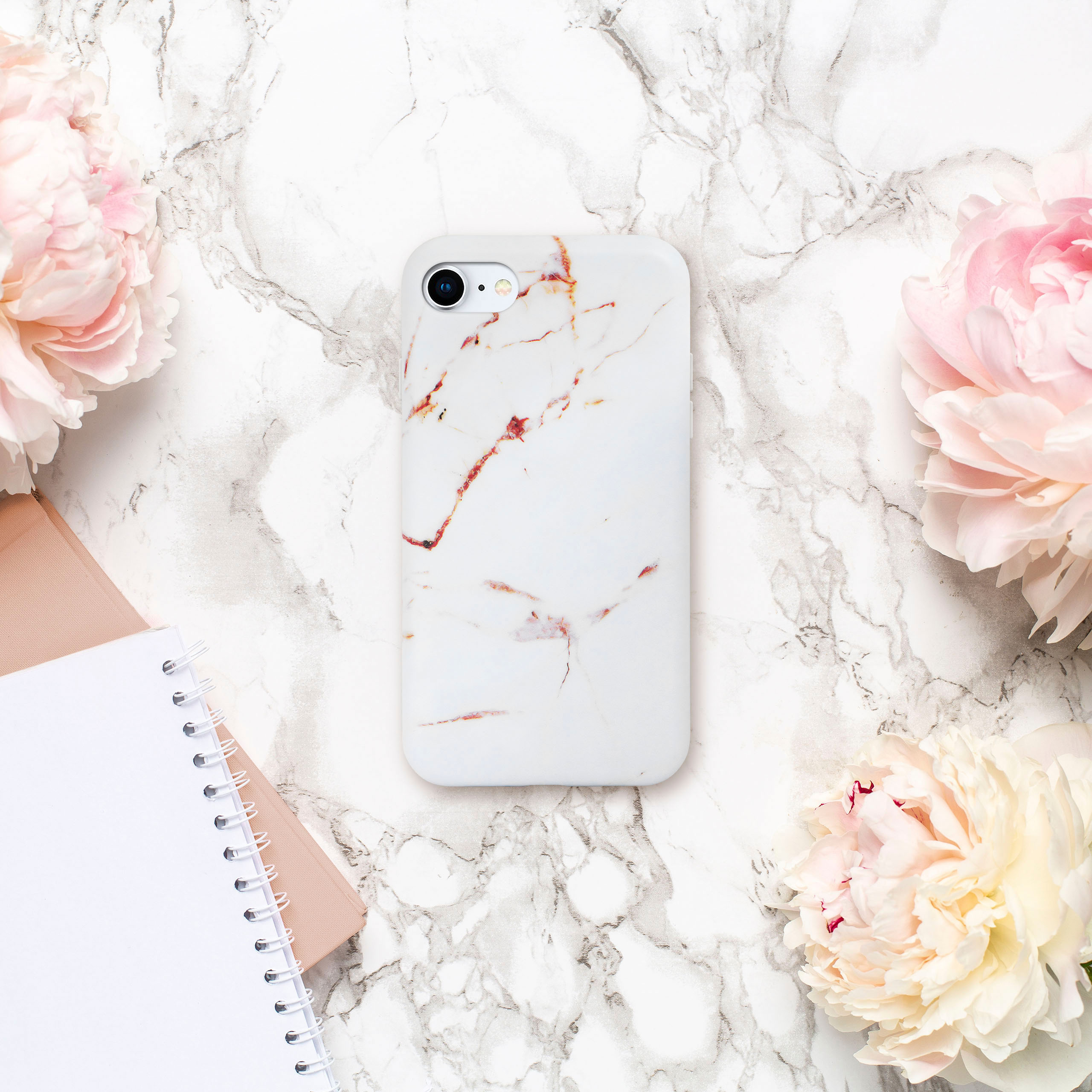 Handyhülle auf Marmoroberfläche mit Blumen und Notizbüchern. Weiße und rosa Farben dominieren.