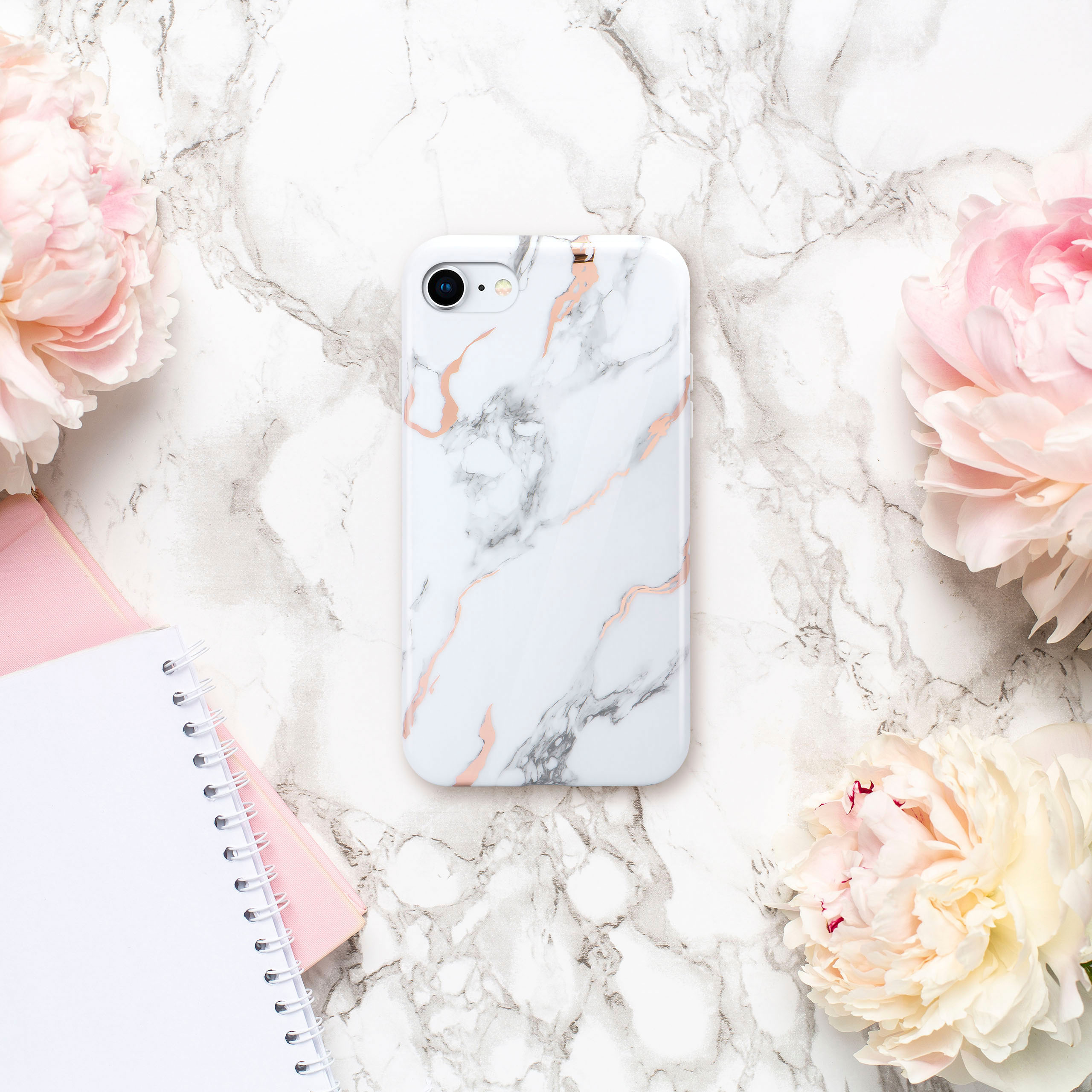 Weißes Marmor-Handycase mit rosa Akzenten liegt auf Marmor, umgeben von Pfingstrosen und einem Notizbuch.