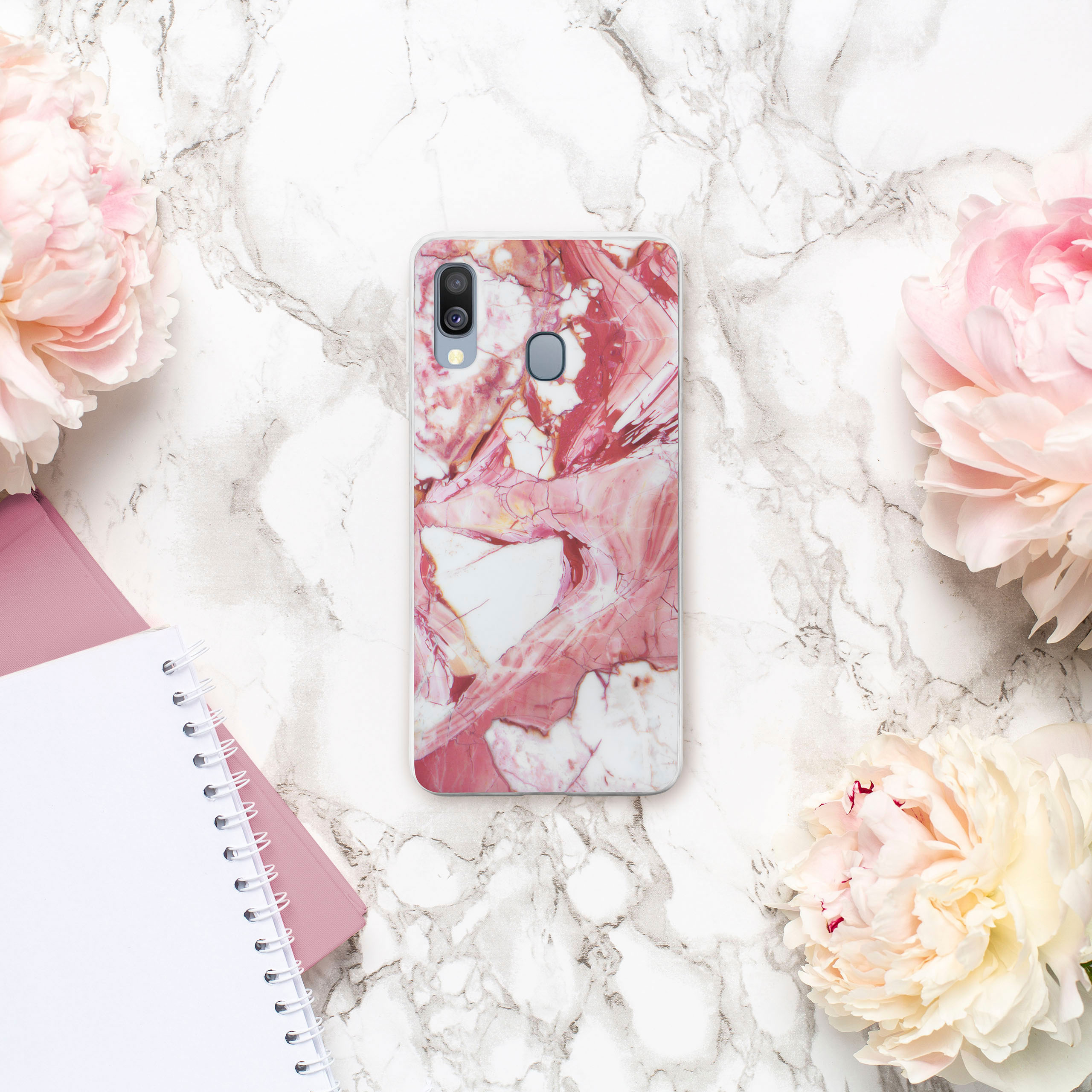 Telefon in einer Hülle mit Marmormuster, auf Marmorhintergrund, umgeben von rosa Blumen und einem Notizblock.