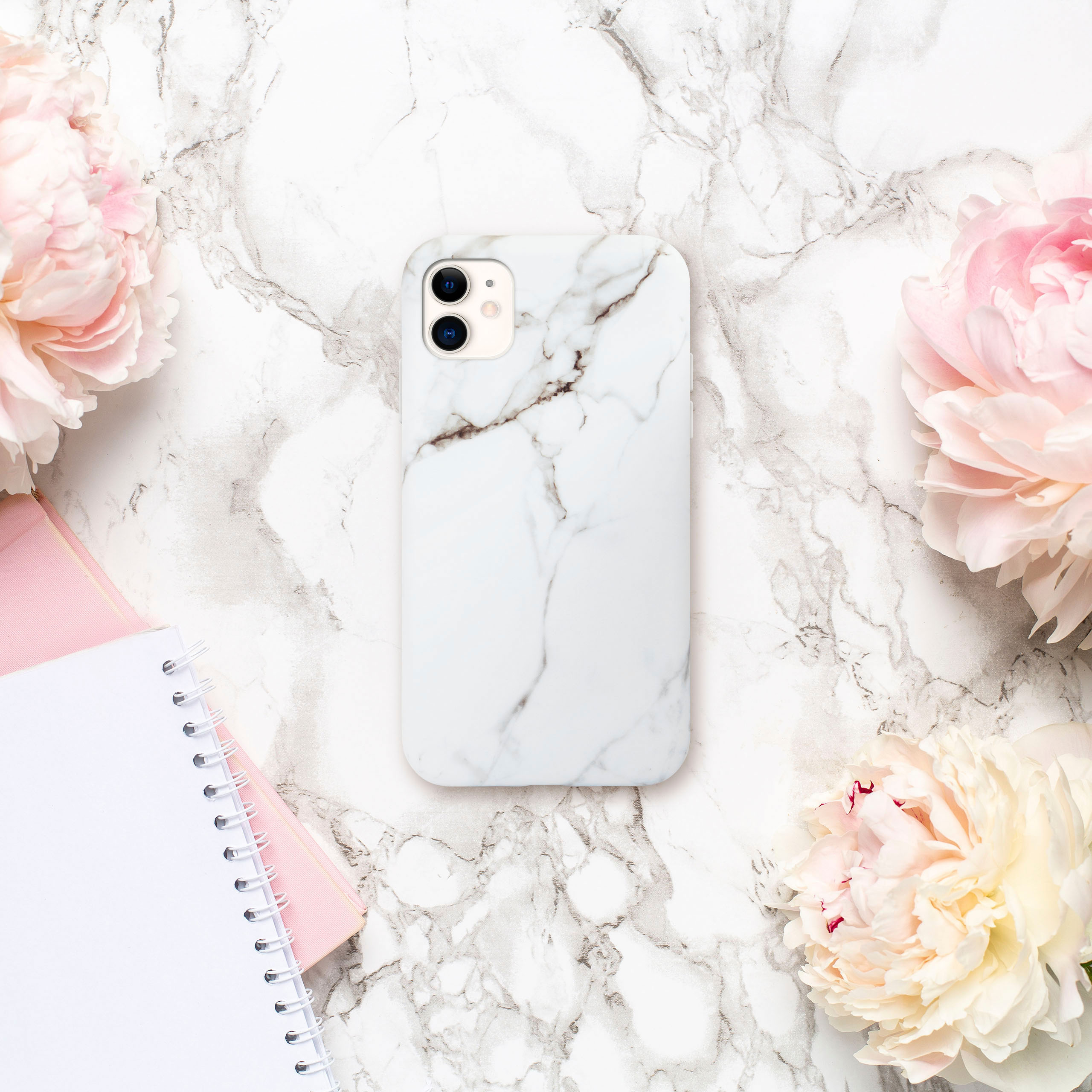 Weißes Marmor-Handycase, Pfingstrosen und ein Notizbuch auf einer Marmoroberfläche.