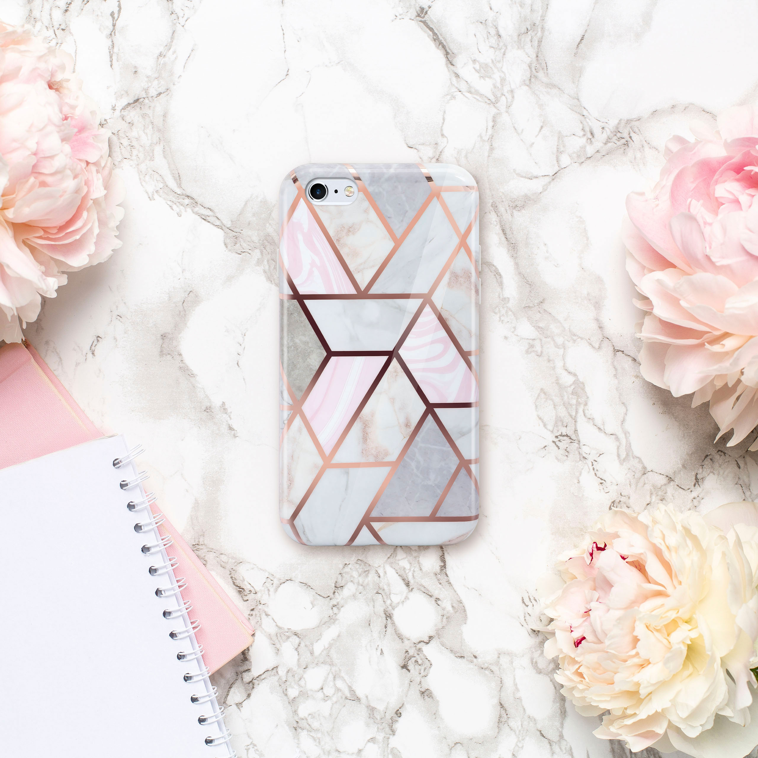 Eine Handyhülle mit geometrischem Marmordesign, umgeben von rosa Blumen und Notizbüchern auf einer Marmoroberfläche.