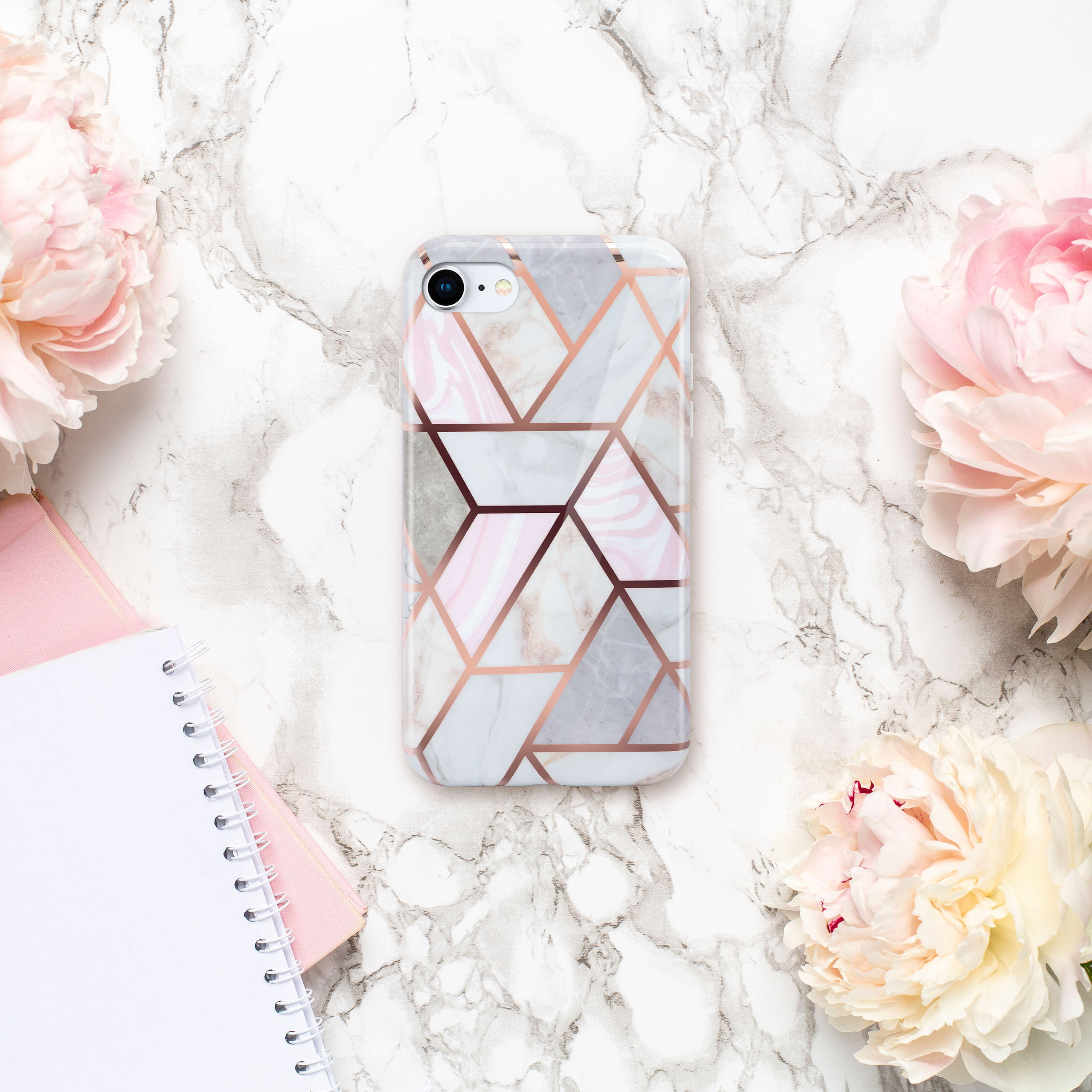 Handyhülle auf Marmoroberfläche mit rosa Pfingstrosen und Notizbüchern, mit geometrischem Roségold-Design.