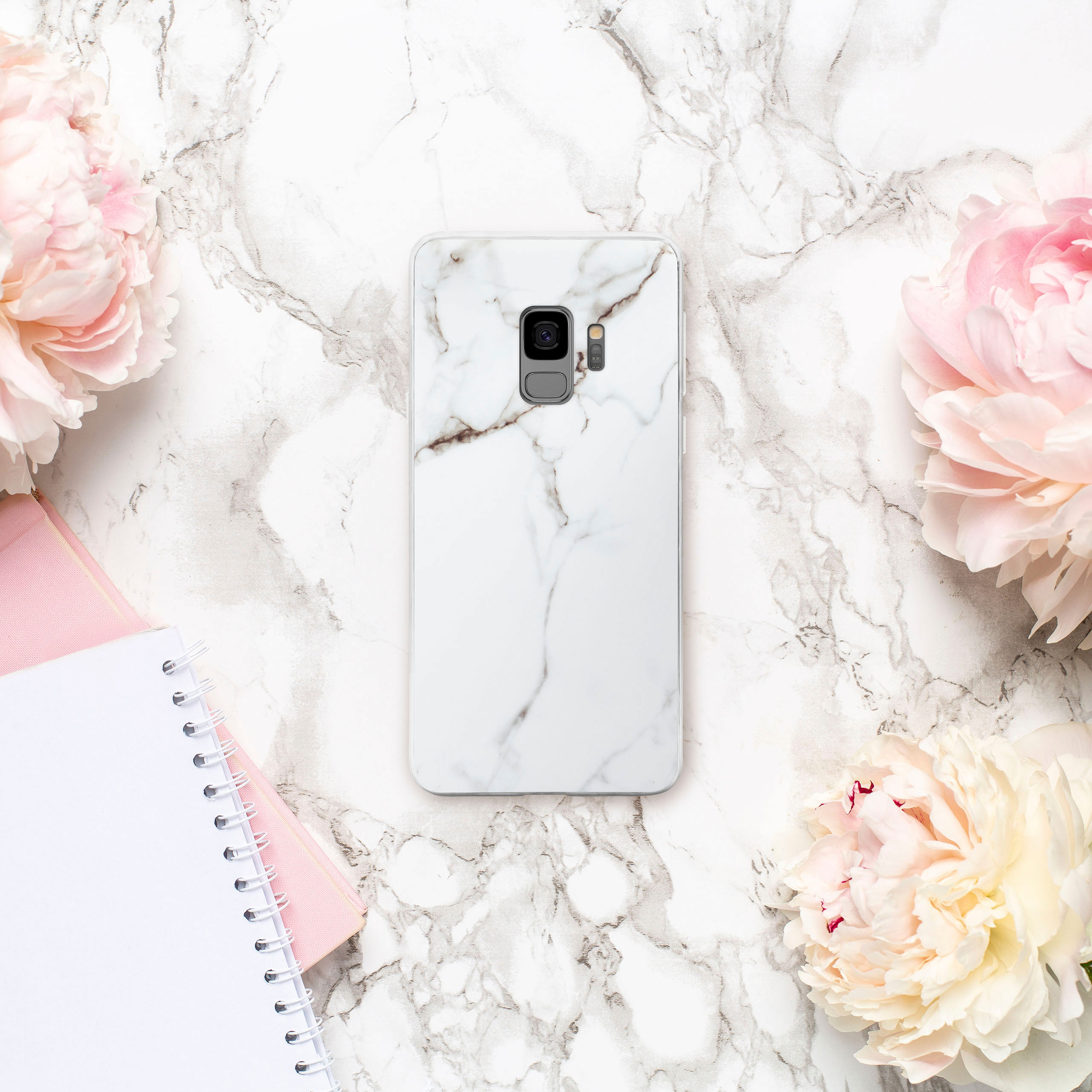 Handyhülle mit Marmordesign, weißer Hintergrund. Rosa Blumen und Notizbuch an der Seite.