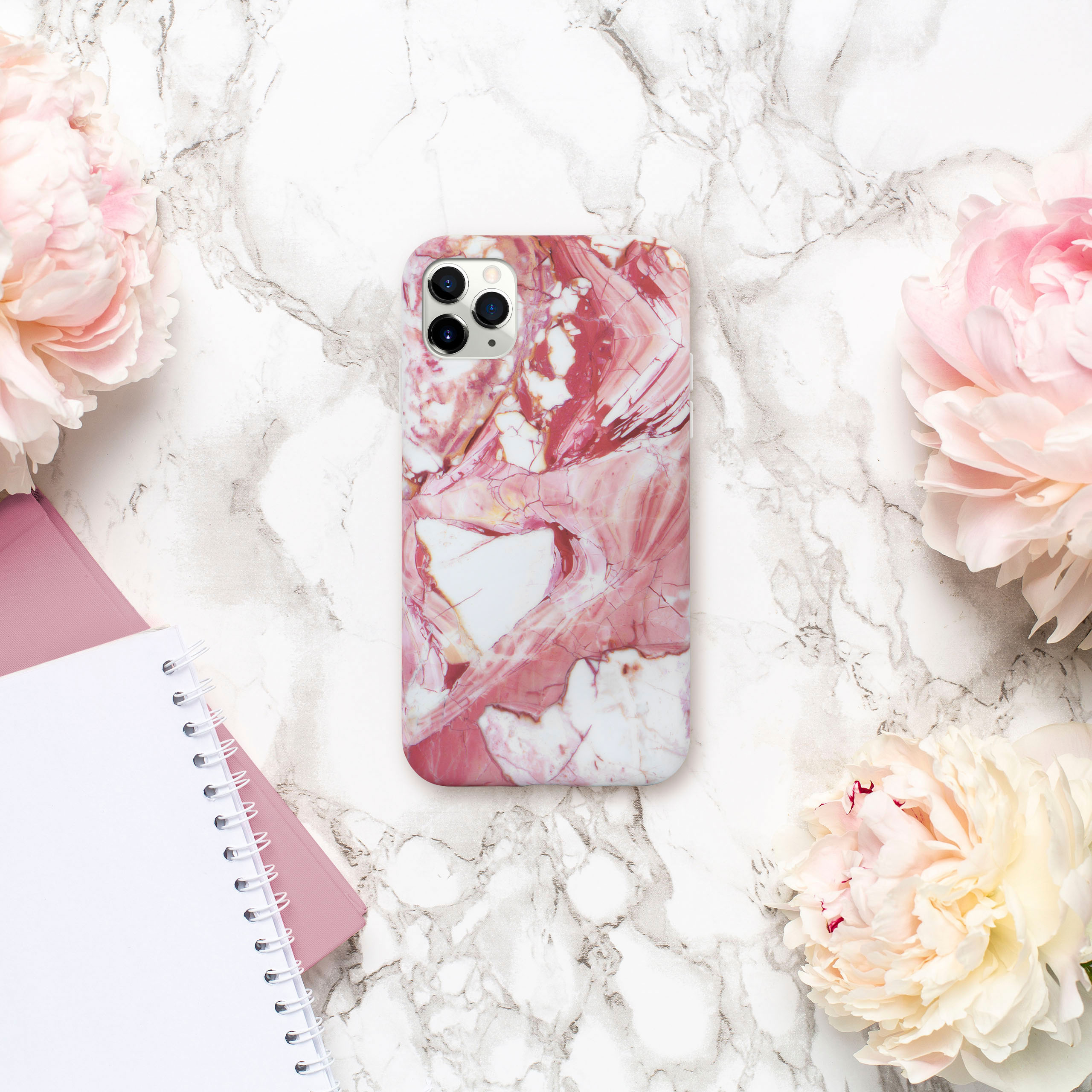 Handyhülle mit rosa Marmordesign, auf Marmoroberfläche platziert. Rosa Blumen und Notizbuch sind darum herum.