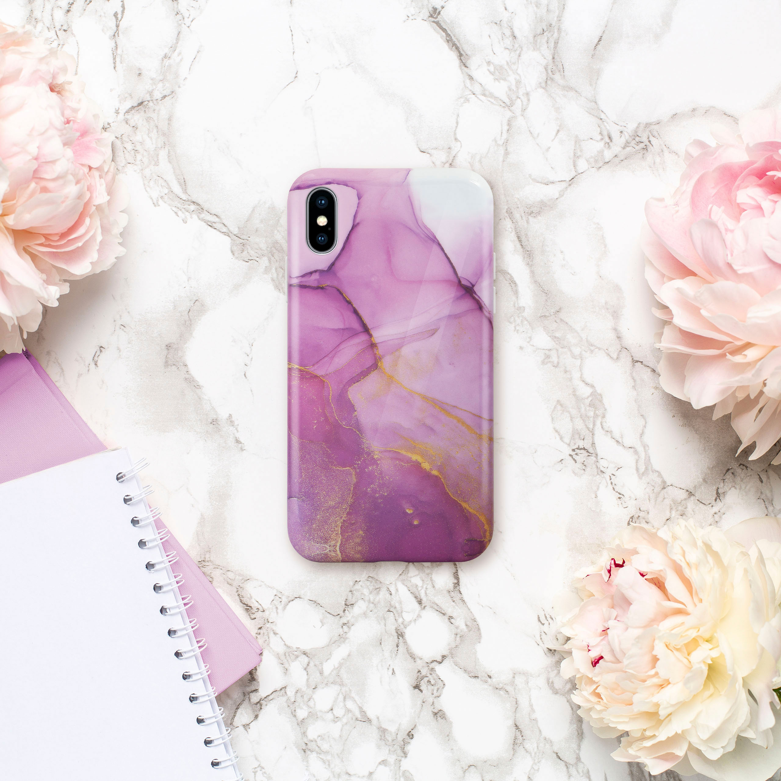 Handyhülle mit lila und goldenem Marmordesign auf einer Marmoroberfläche mit rosa Pfingstrosen und Notizbüchern.