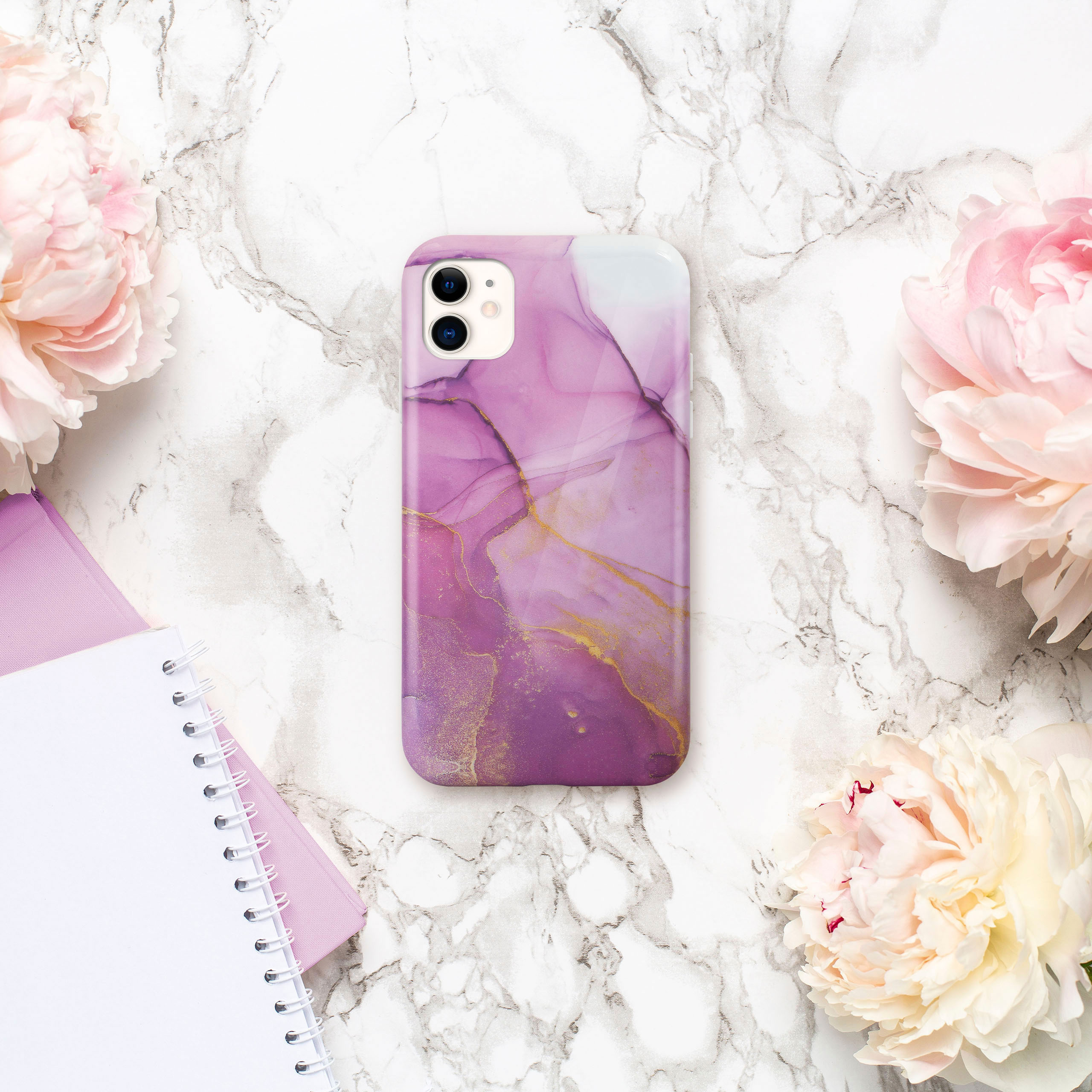 Handyhülle auf Marmor, mit lila und goldenem abstraktem Design, umgeben von rosa Pfingstrosen und Notizbüchern.