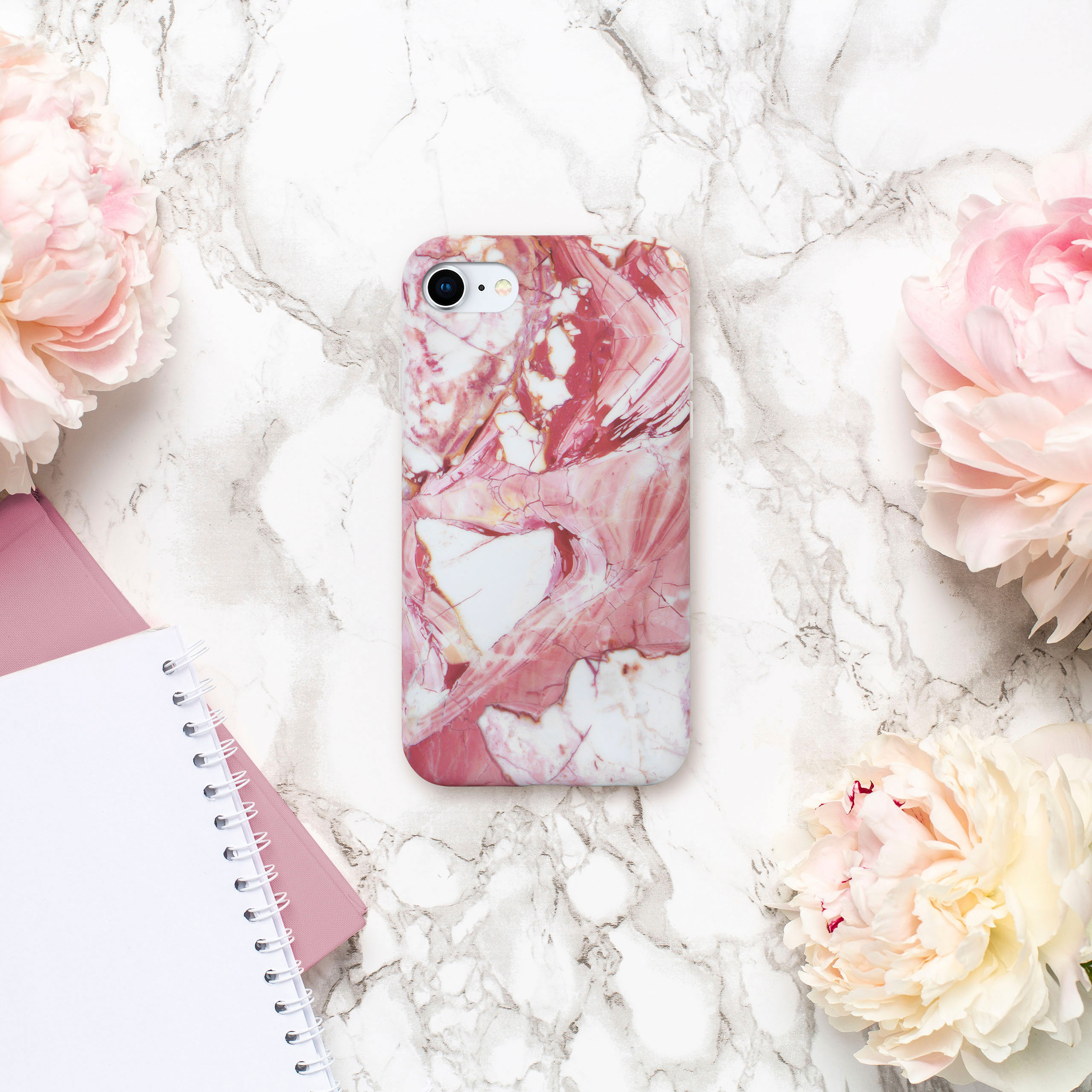 Handyhülle mit Marmordesign, rosa Pfingstrosen und einem Notizbuch auf weißer Marmoroberfläche.