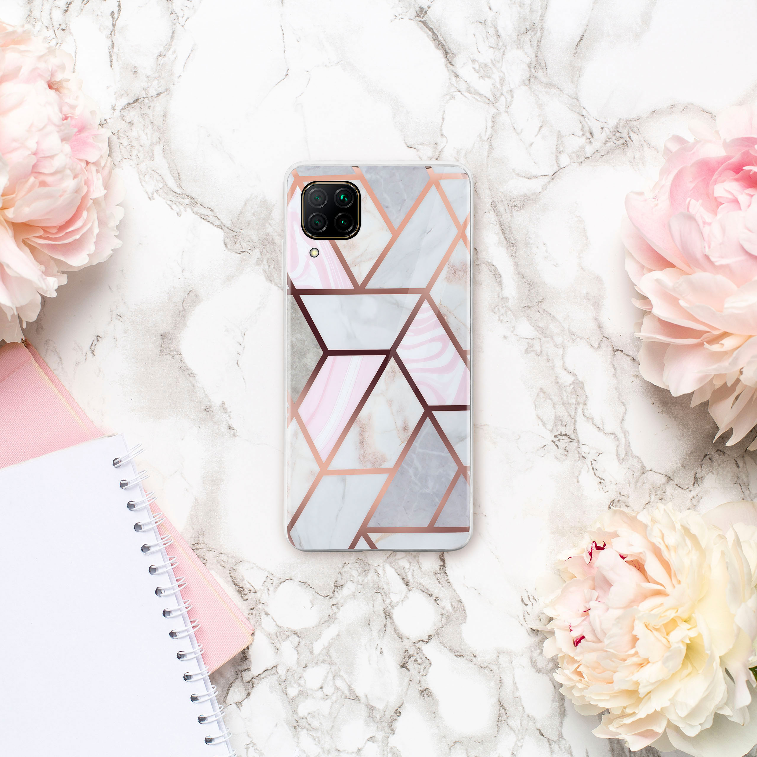 Handyhülle mit geometrischem Design auf Marmoroberfläche. Rosa Pfingstrosen und Notizbuch sichtbar.