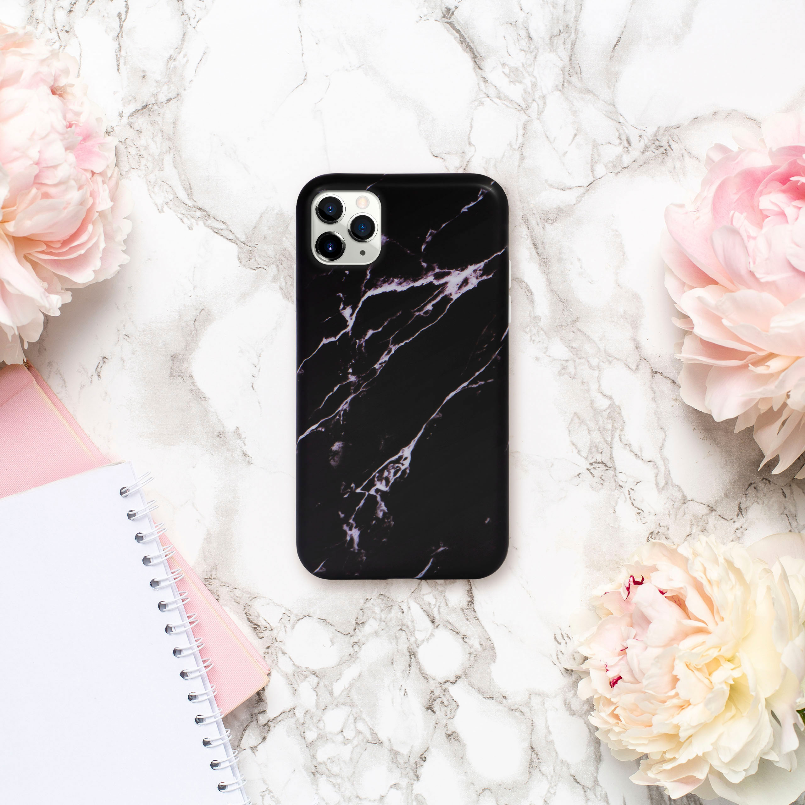 Handyhülle mit schwarzem Marmordesign auf Marmorhintergrund, umgeben von rosa Blumen und Notizbüchern.
