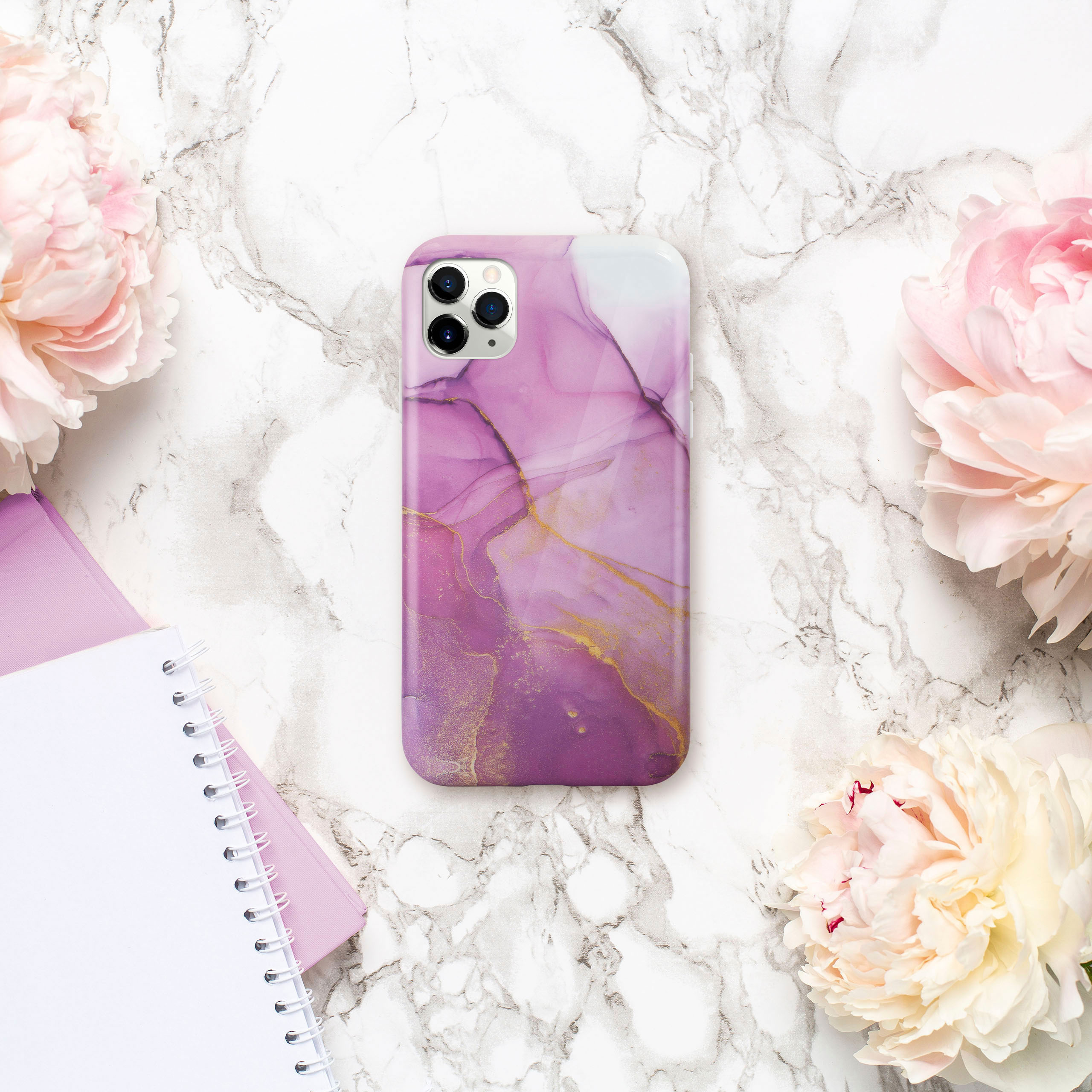 Handyhülle mit Marmordesign, rosa und gold. Umgeben von Pfingstrosen und einem Notizbuch auf Marmoroberfläche.
