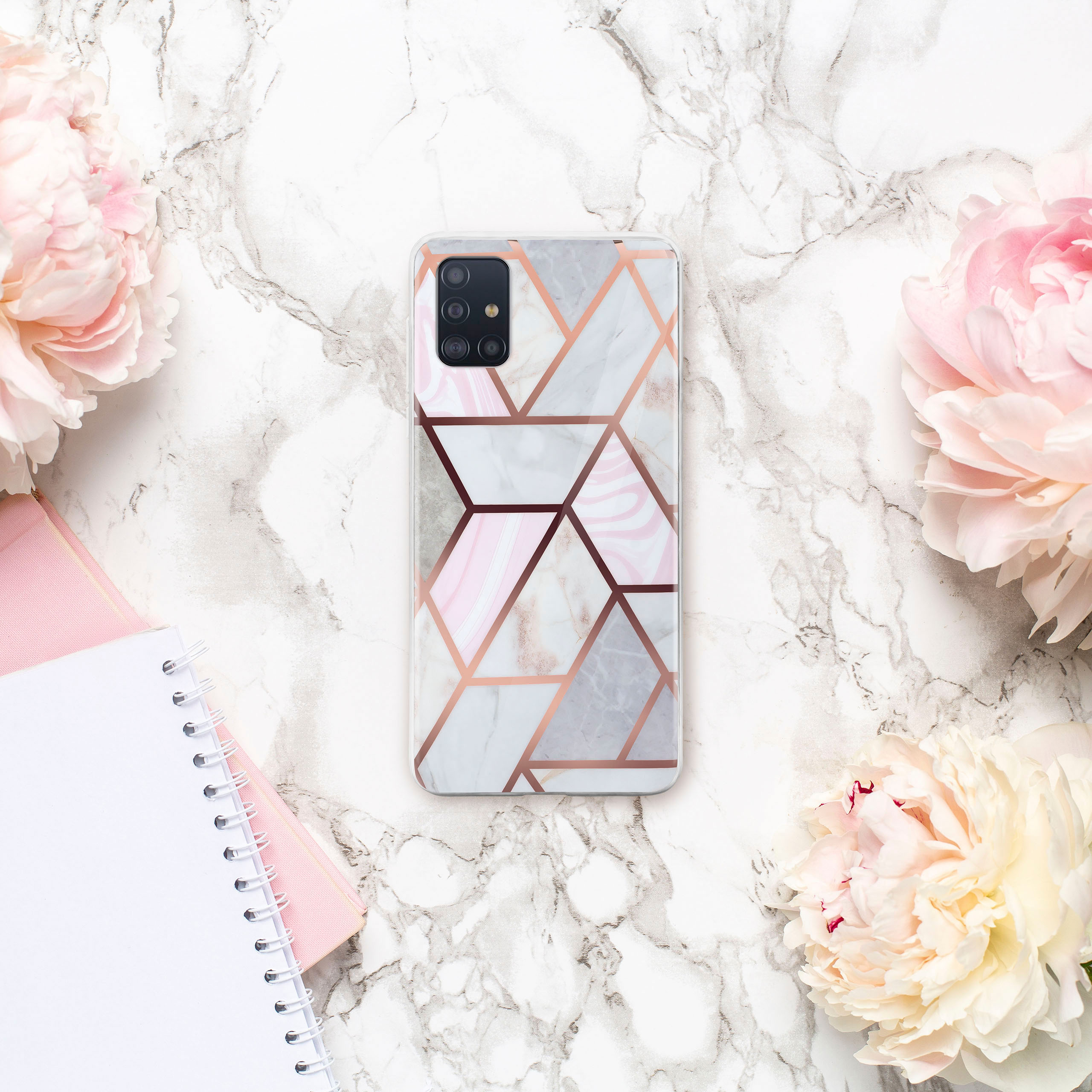 Handyhülle mit Marmor- und rosa geometrischem Design, umgeben von Blumen und Notizbüchern auf Marmorhintergrund.