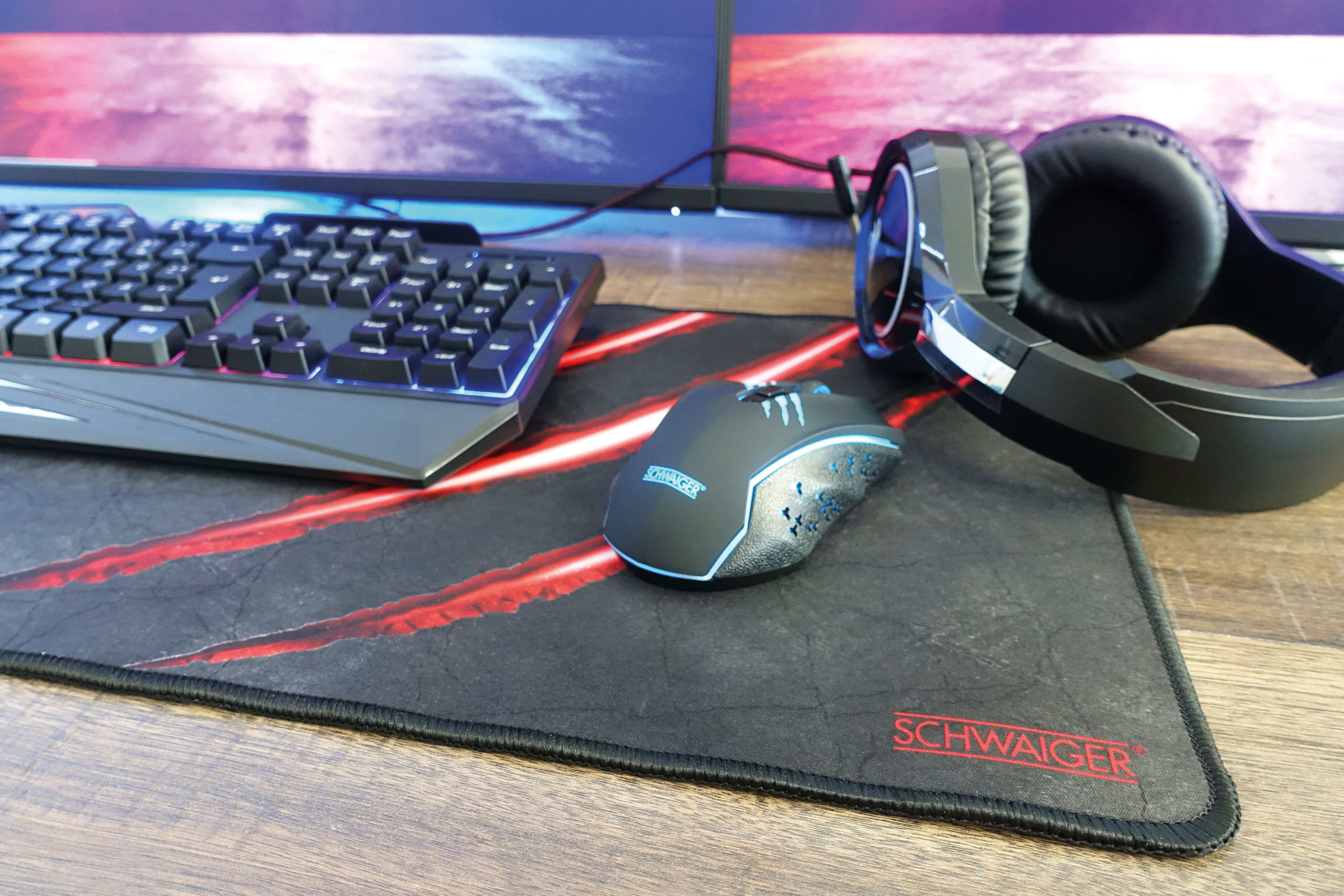 Gaming-Setup: Tastatur, Maus, Mauspad und Kopfhörer. Zwei Monitore im Hintergrund.