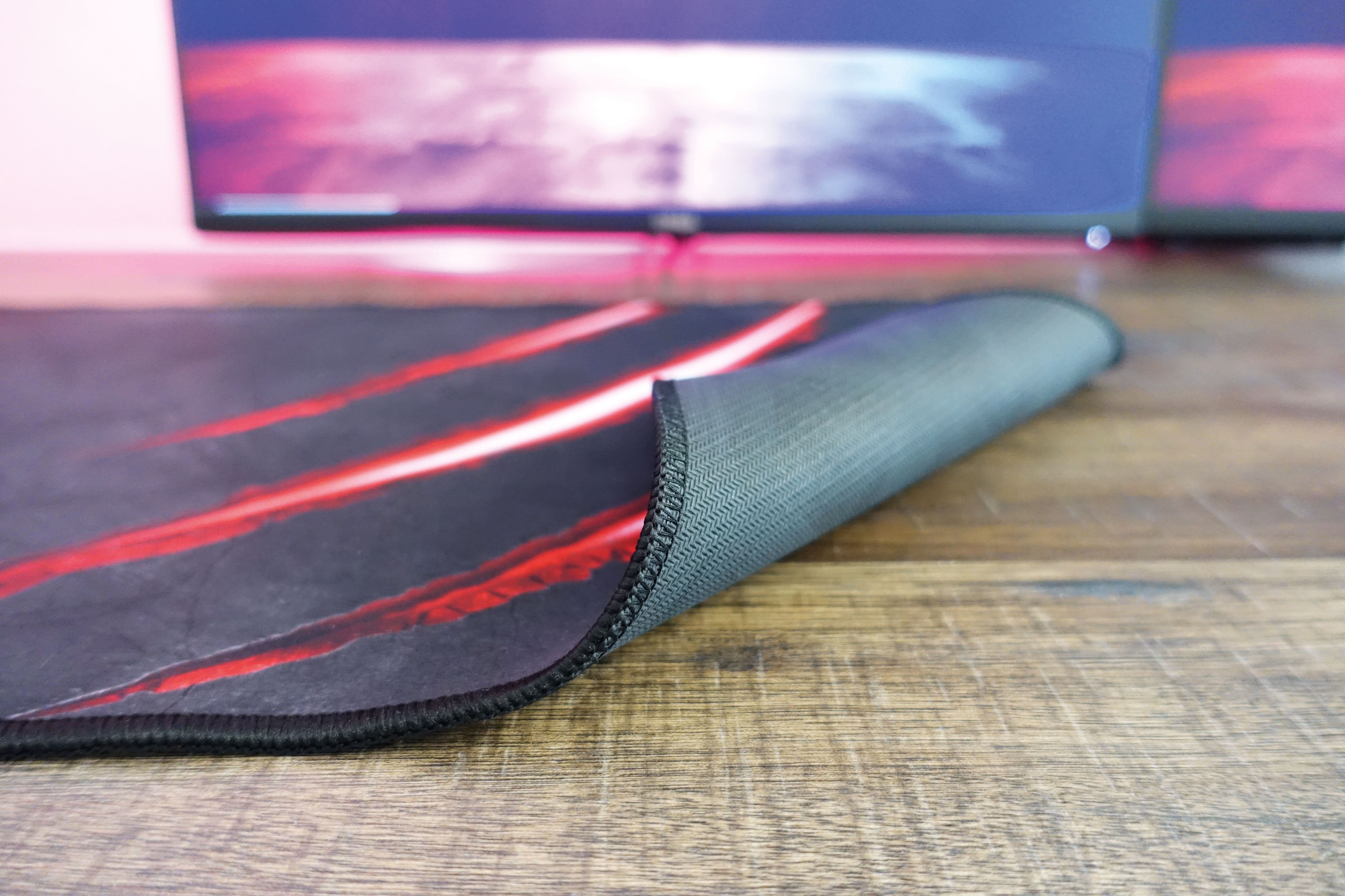 Ein Mousepad mit rotem Design auf einem Holztisch. Im Hintergrund ist ein Computerbildschirm zu sehen.