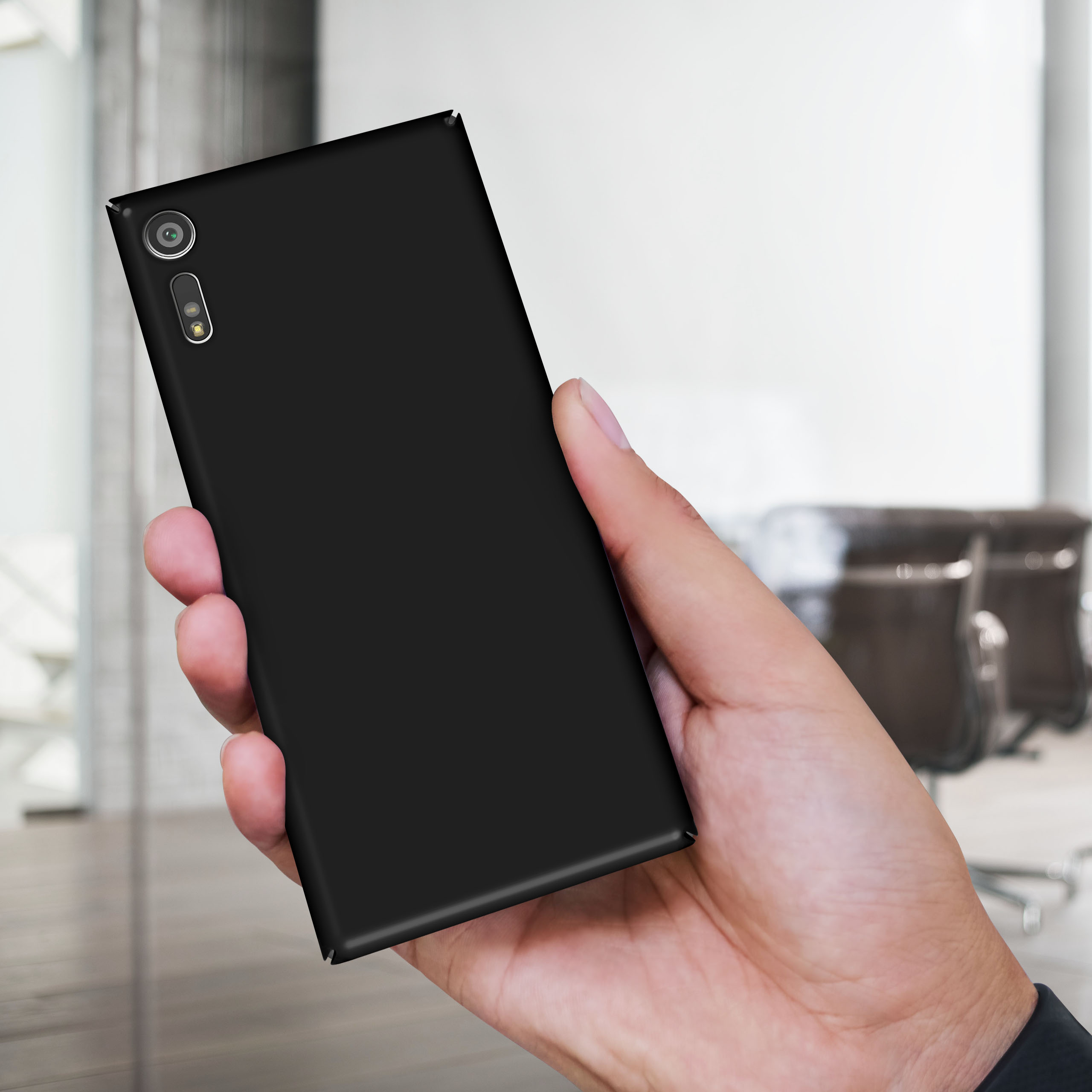 Hand hält ein schwarzes Telefon, die Kamera ist sichtbar. Verschwommenes Büro im Hintergrund.