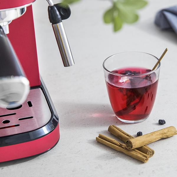 Rote Espressomaschine, ein Glas mit roter Flüssigkeit, Zimtstangen und ein paar schwarzen Samen auf einer Oberfläche.