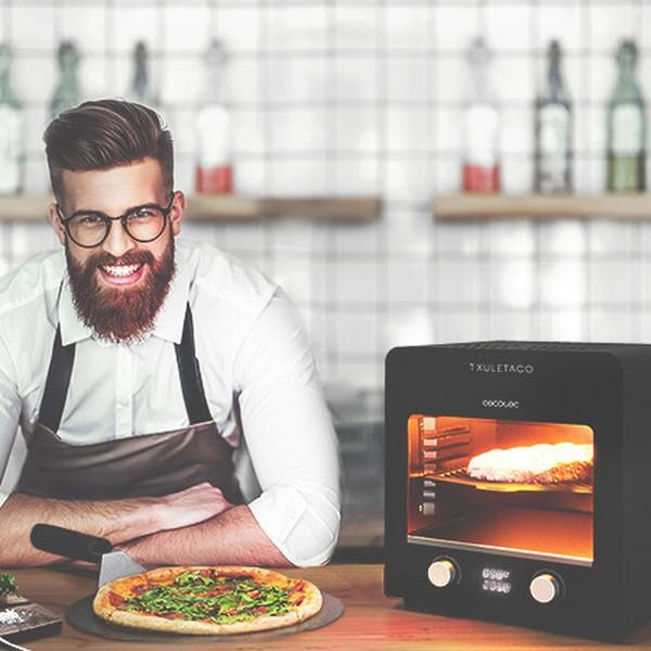 Een man lacht in de buurt van een pizza en een zwarte oven. De achtergrond toont planken.