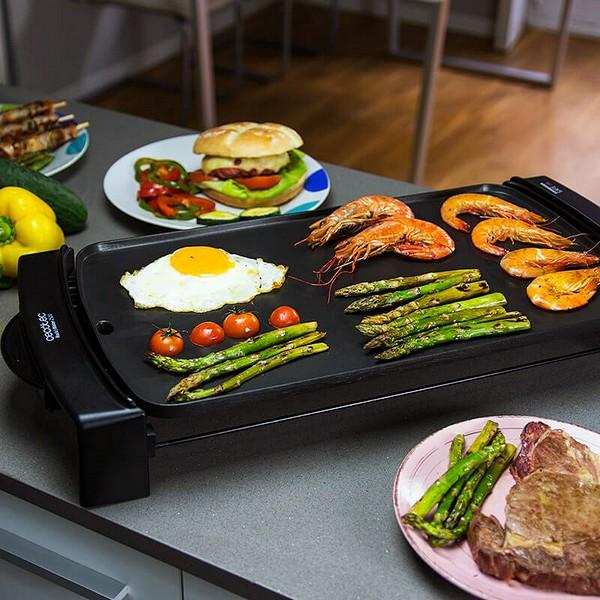 Una piastra nera con vari cibi cotti come gamberi, asparagi, uova e un hamburger su un tavolo.